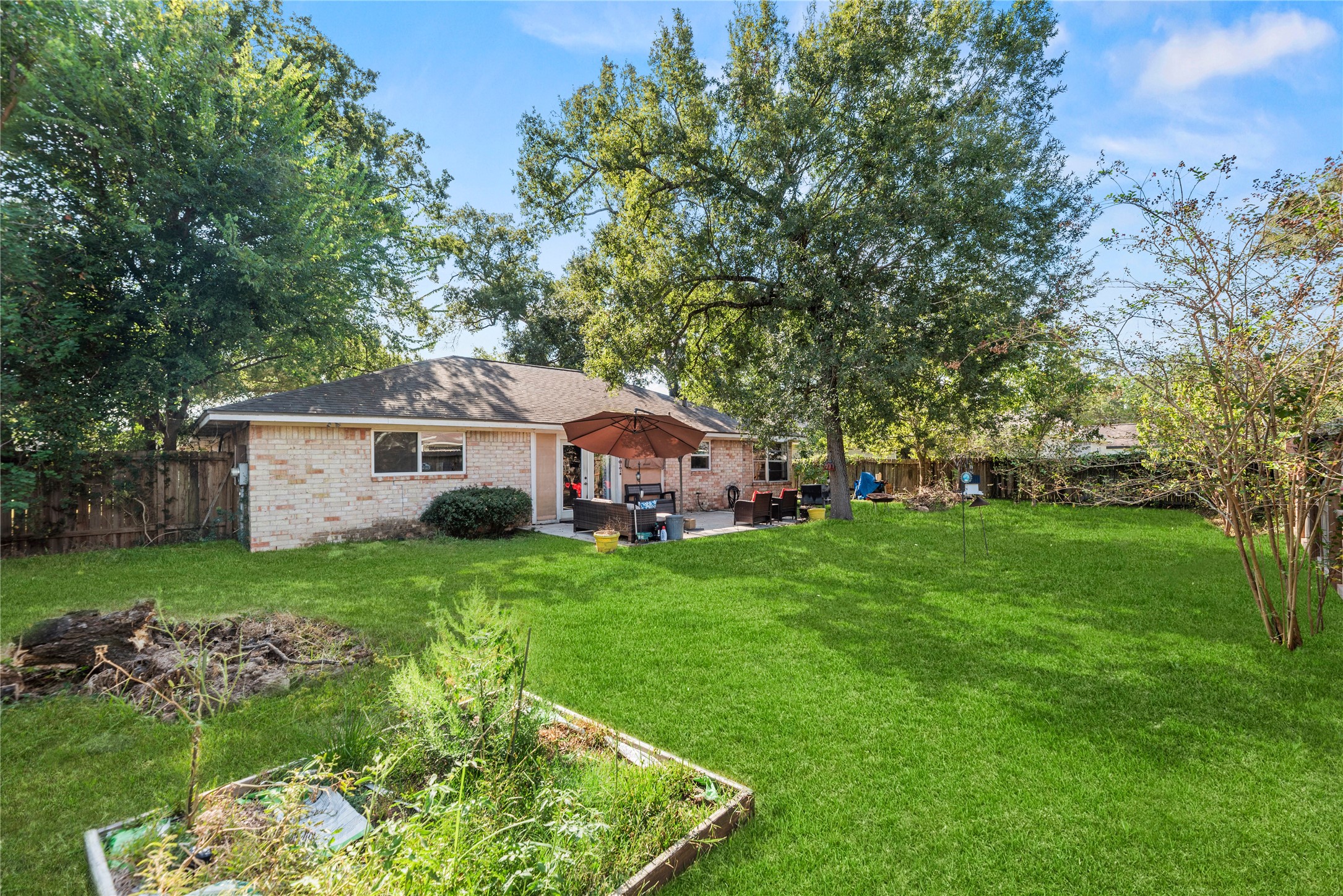 Original backyard view toward the home's brick rear exterior and patio, showing existing garden areas and yard items before virtual clearing.