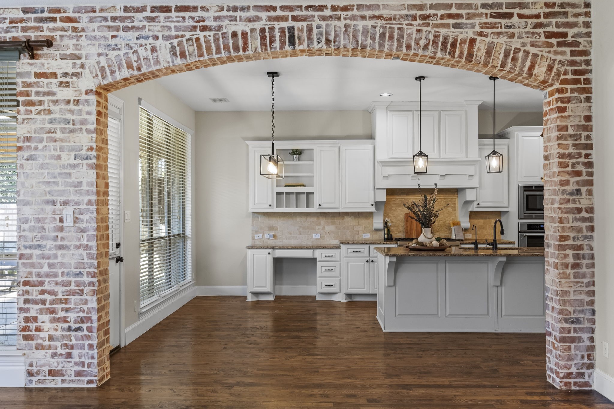 A beautiful brick archway frames the spacious kitchen, featuring white cabinetry, granite countertops, a large center island with seating, pendant lighting, and rich hardwood floors. A charming architectural detail that creates a seamless connection between the kitchen and family room.