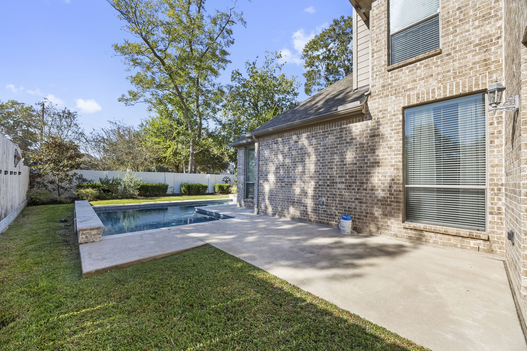 Another view of the backyard highlights the additional concrete patio area, perfect for outdoor dining, grilling, or creating a second seating space. The combination of open lawn, mature trees, and poolside living offers plenty of options for enjoying the outdoors.