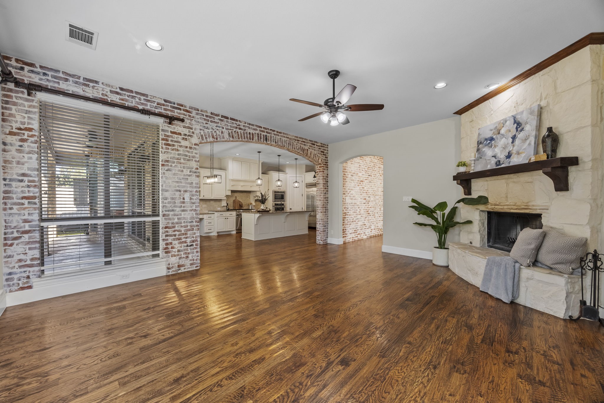 Spacious family room featuring rich hardwood floors, a stunning floor to ceiling stone fireplace, and beautiful brick accent walls with an arched opening to the kitchen. A warm, inviting space perfect for everyday living and entertaining.