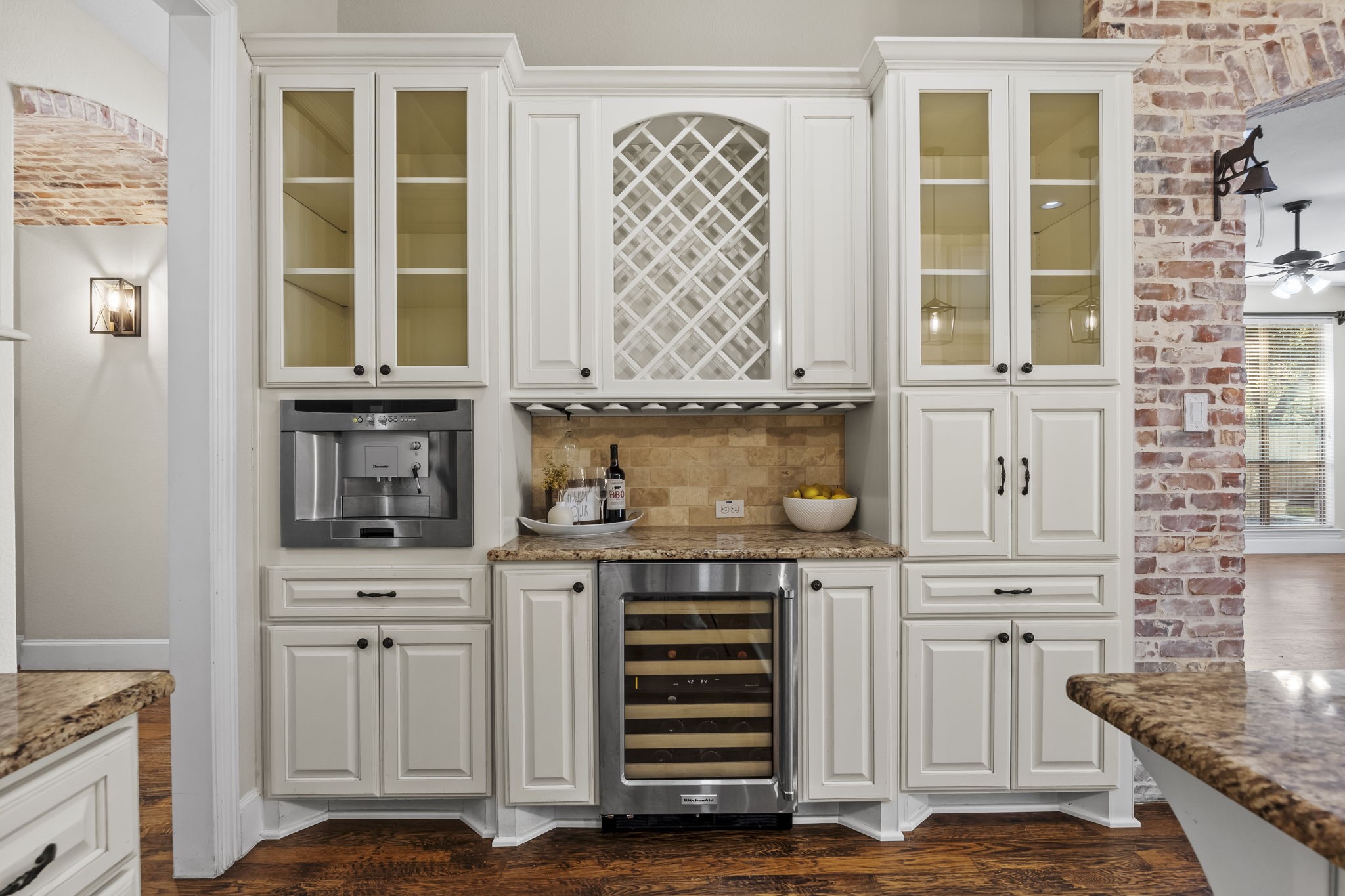 Beautifully designed beverage station featuring custom white cabinetry, glass front display cabinets, decorative wine storage, granite countertops, a built-in wine refrigerator, and integrated coffee machine. A perfect space for entertaining and keeping your favorite beverages close at hand