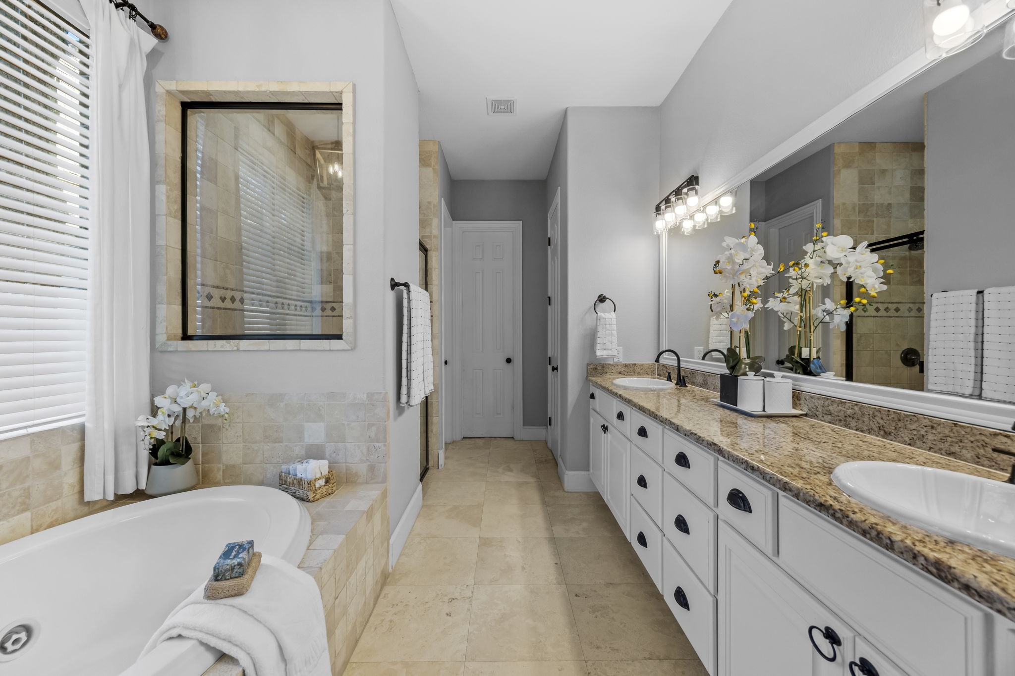 The primary bathroom features a long dual sink vanity with abundant storage, a jetted soaking tub, and a separate glass enclosed shower. Natural light and neutral tile finishes create a relaxing, spa inspired retreat.