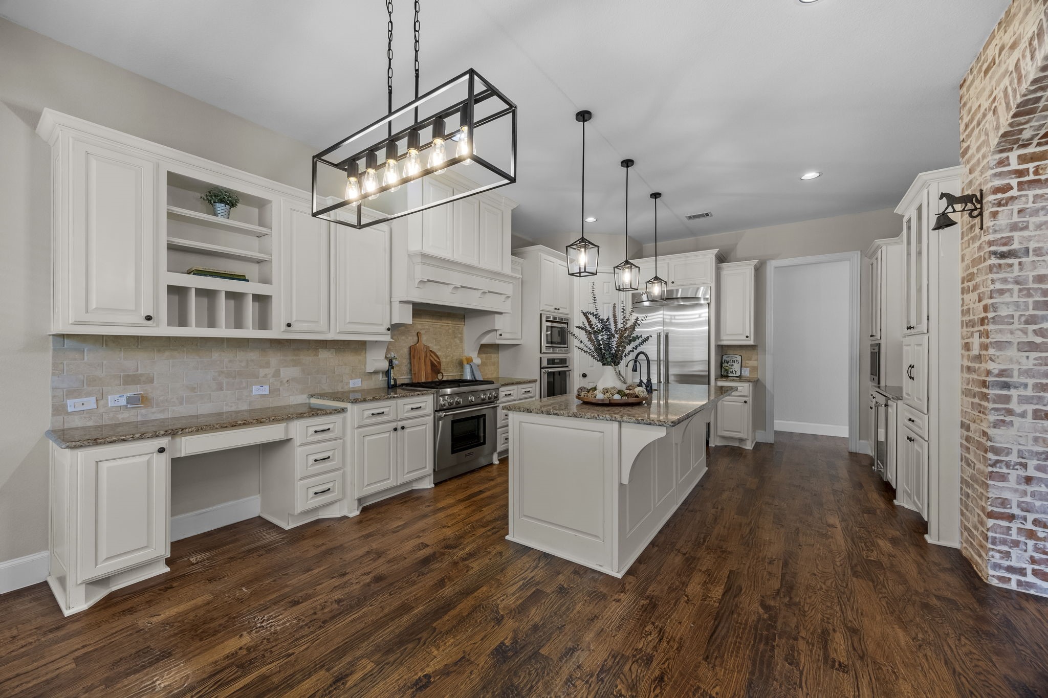 chef's kitchen featuring beautiful white cabinetry, granite countertops, stainless steel appliances, and a large center island with sink. Custom vent hood, built-in desk area, decorative pendant lighting, rich hardwood floors, and charming brick accents add character and functionality to this inviting space.