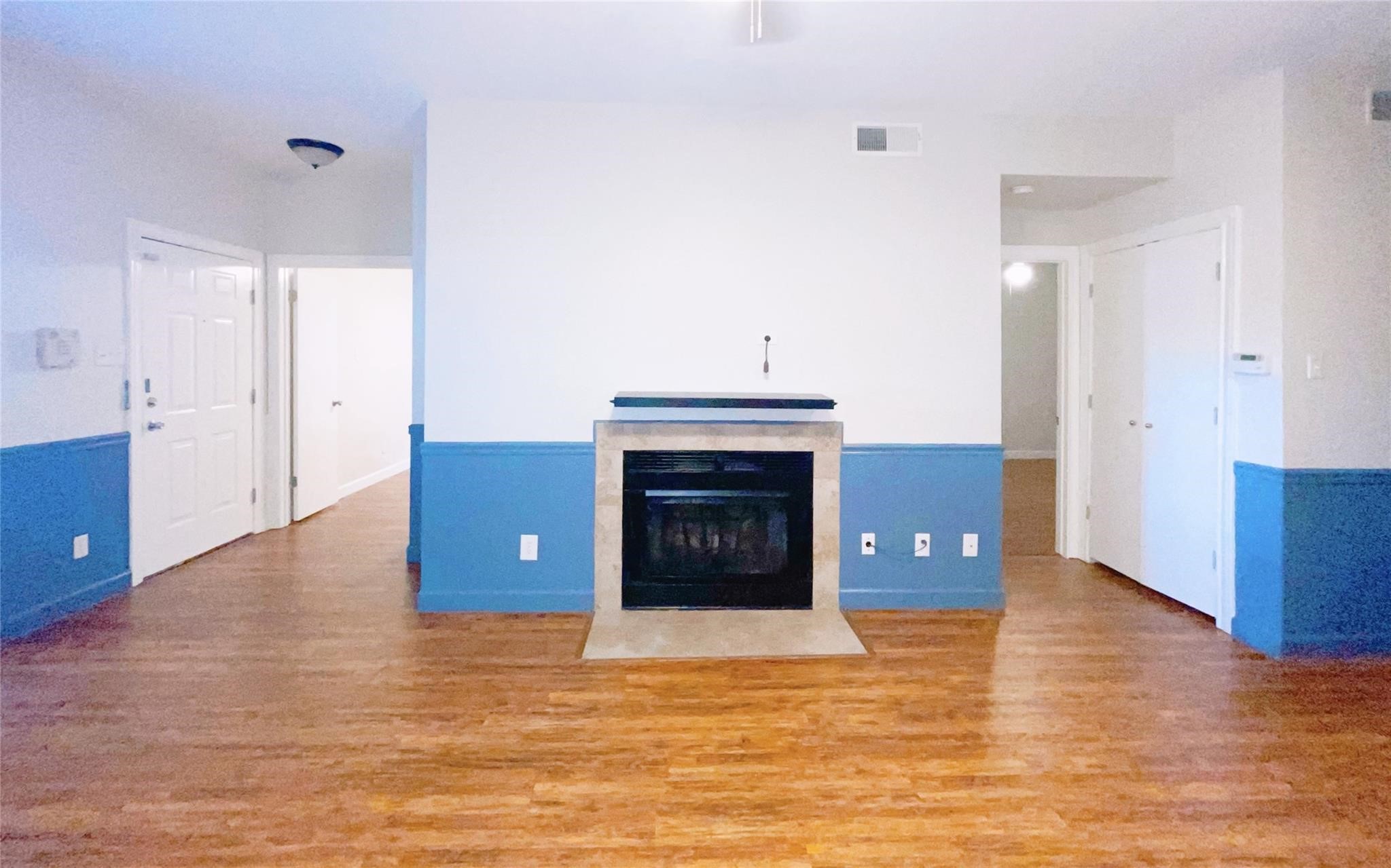 Spacious living area featuring warm wood floors, a fireplace with stone surround, and attractive blue gray wainscoting. The open layout provides a comfortable gathering space with convenient access to the adjoining area of the home.