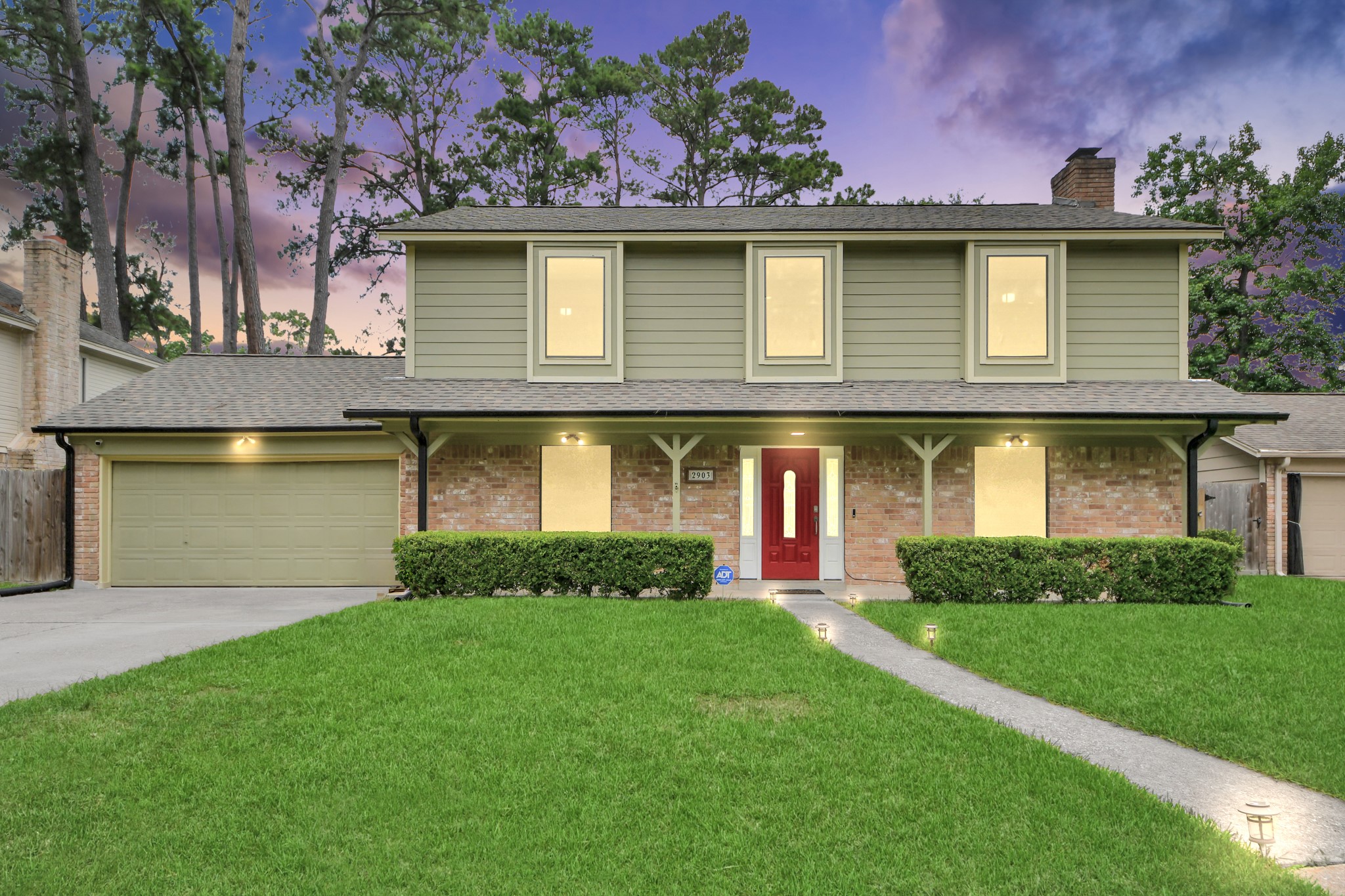 An enchanting evening welcome. Captured at twilight, the glowing windows cast a warm radiance across the classic brick and cement board facade, accented by a striking red front door and a beautifully manicured, expansive lawn.