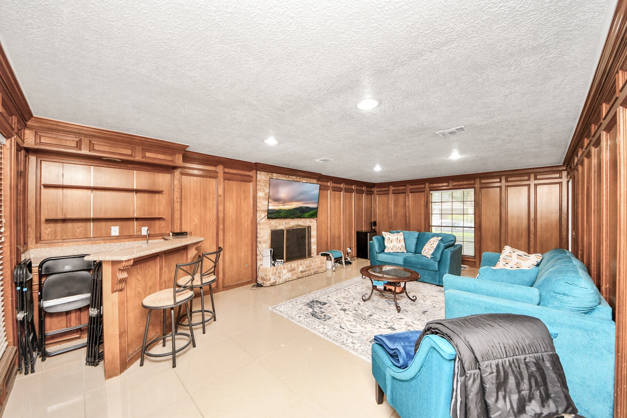 Mid-century warmth meets modern luxury. Gorgeous, rich wood paneling anchors this magnificent living room, dynamically equipped with a dedicated wet bar, a central fireplace, and oversized windows.