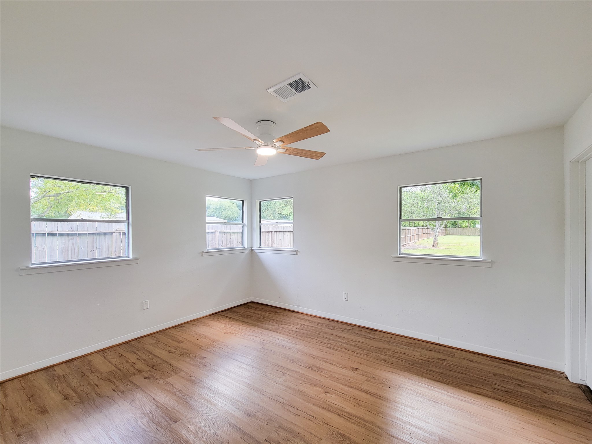 A bright and airy room featuring large windows, beautiful wood flooring, and a modern ceiling fan, all offering a serene view of the backyard.
