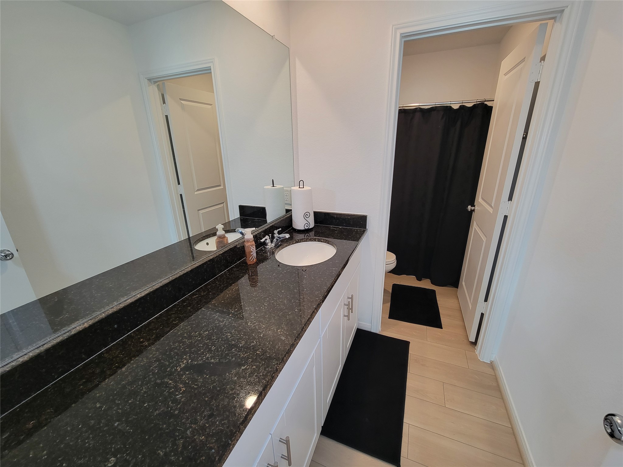 This bathroom features a sleek black granite countertop with dual sinks and modern fixtures. It includes a large mirror, light wood flooring, and a separate area with a shower and toilet, enhancing privacy.