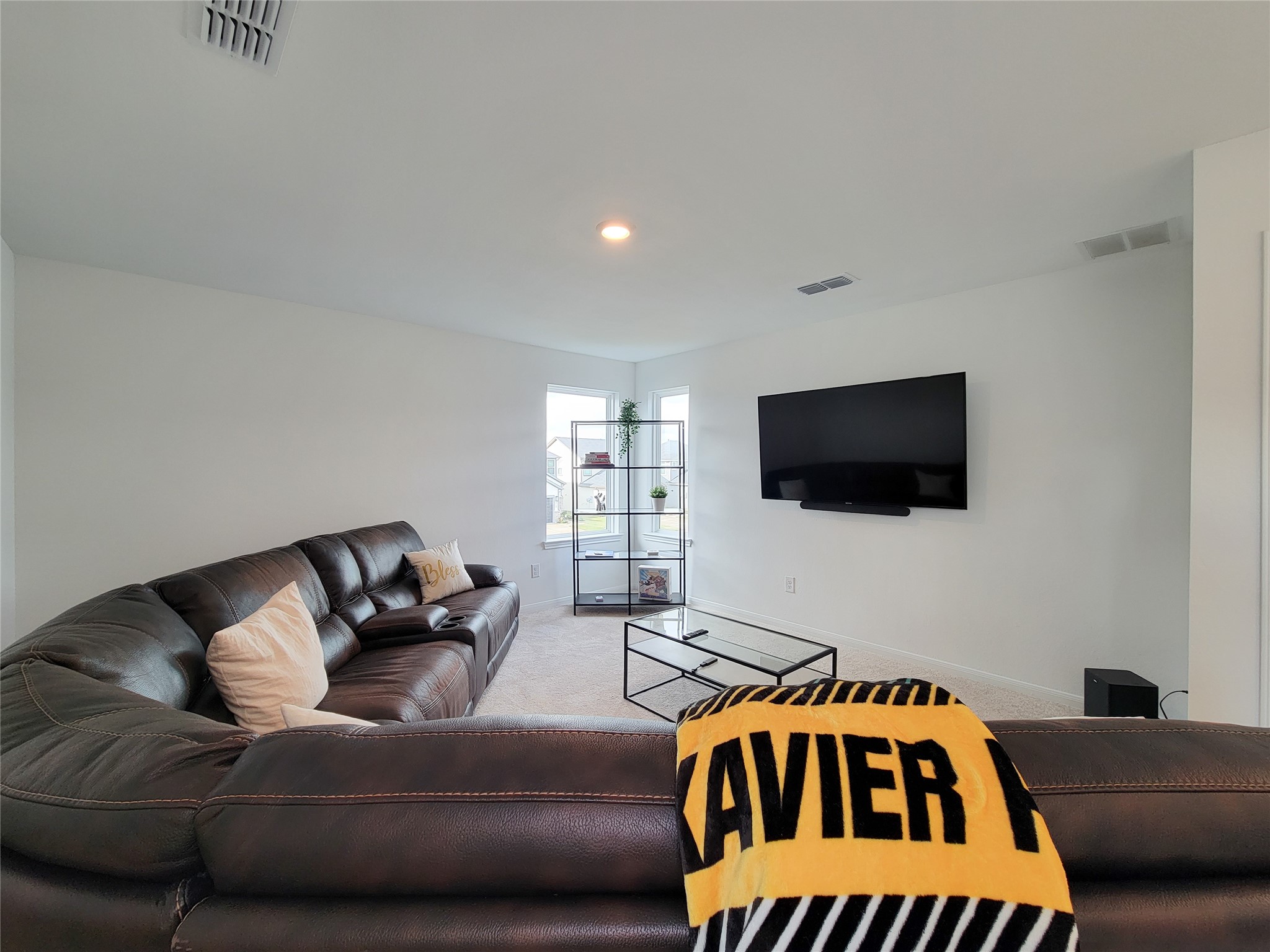 This spacious living room features a cozy, dark leather sectional, a wall-mounted TV, and a modern glass coffee table. A window brings in natural light, enhancing the room's airy feel. Perfect for relaxation and entertainment.