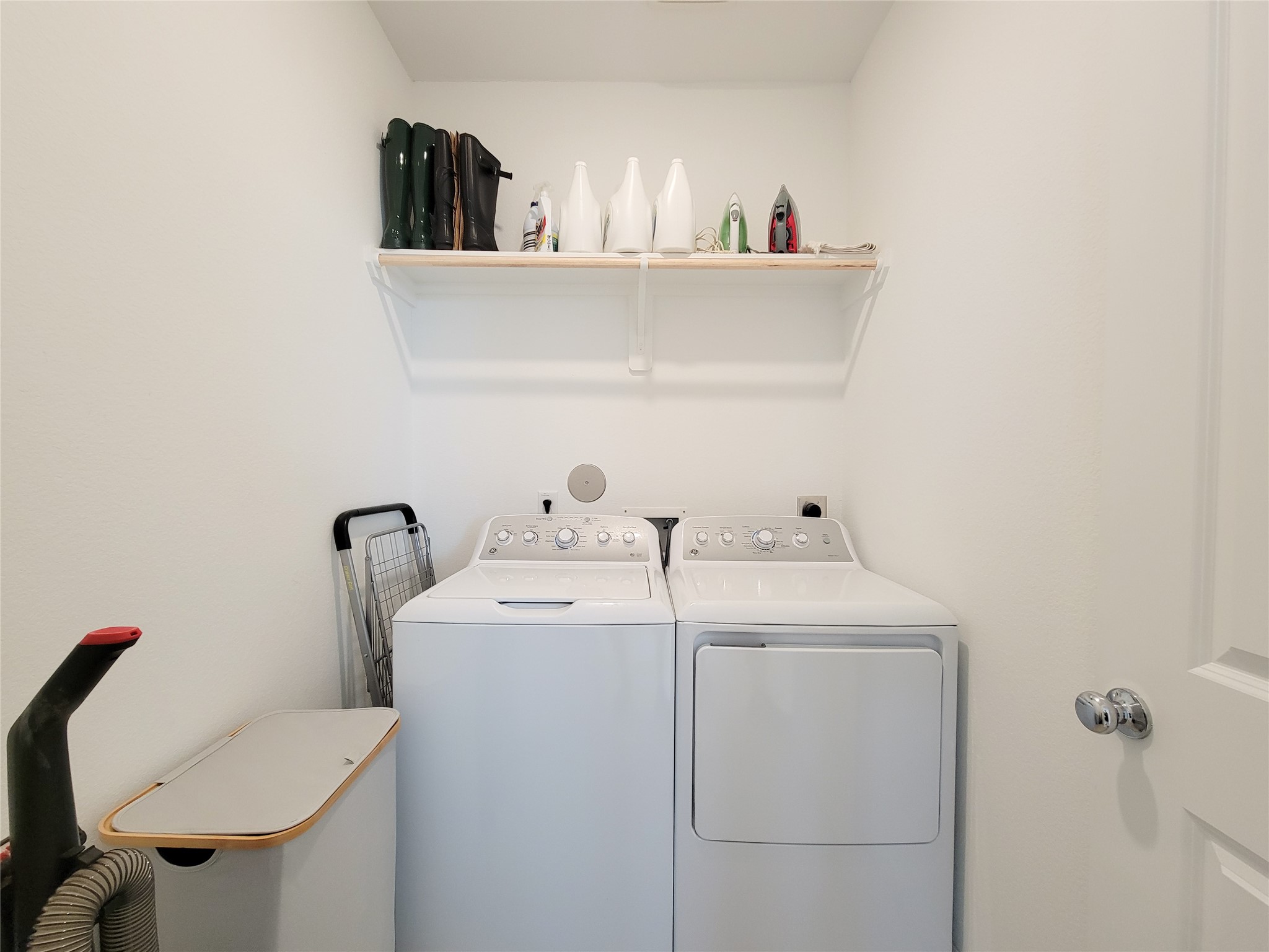 Compact laundry room with a washer and dryer set. It includes a shelf above for storage, holding items like detergent and an iron. The space is well-organized and has a clean, white finish.