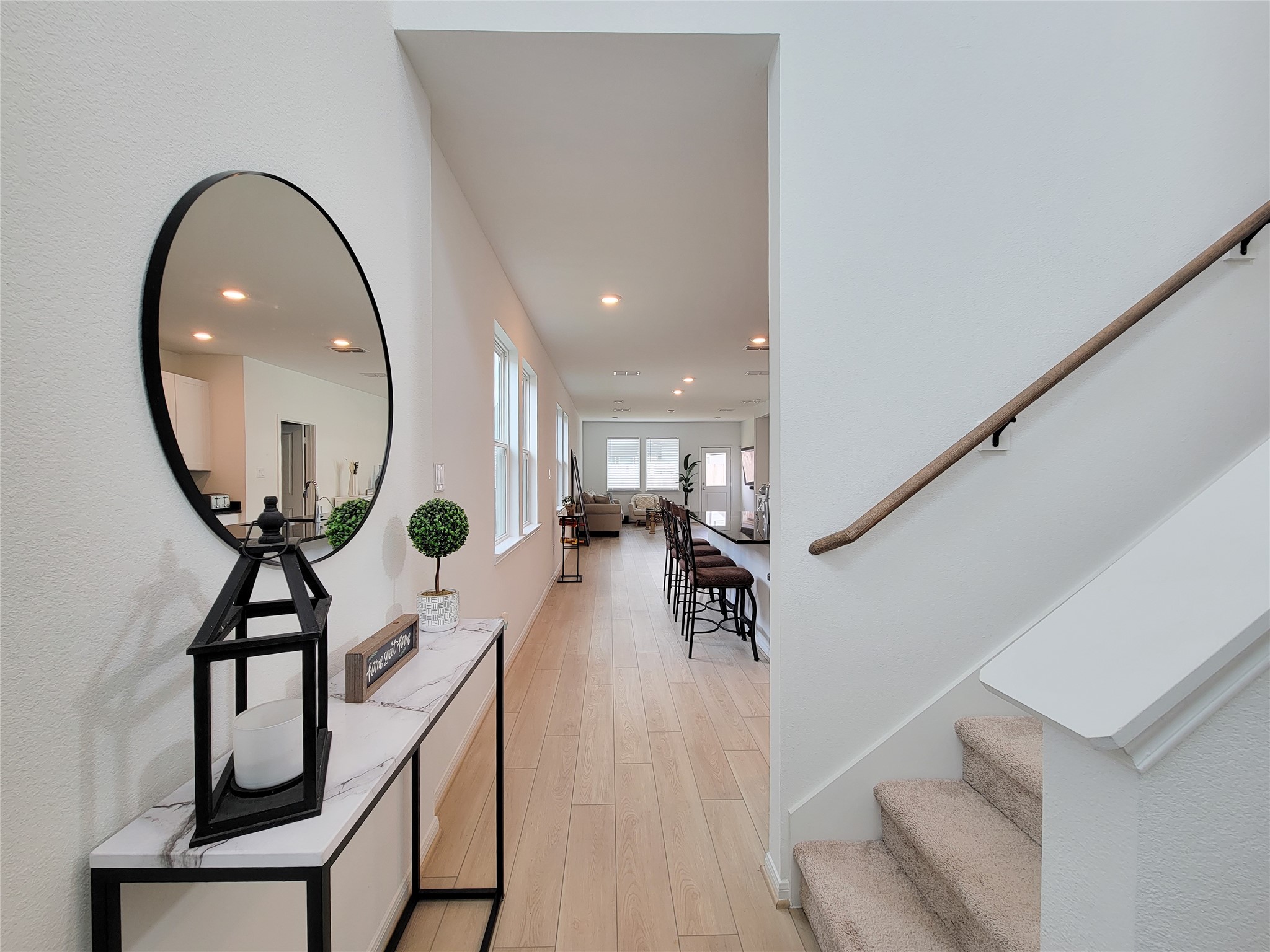 Entryway with modern decor, featuring a large round mirror and a sleek console table. The space leads into an open-plan living area with wooden flooring, ample natural light, and a glimpse of the dining and living rooms. A carpeted staircase is visible on the right.