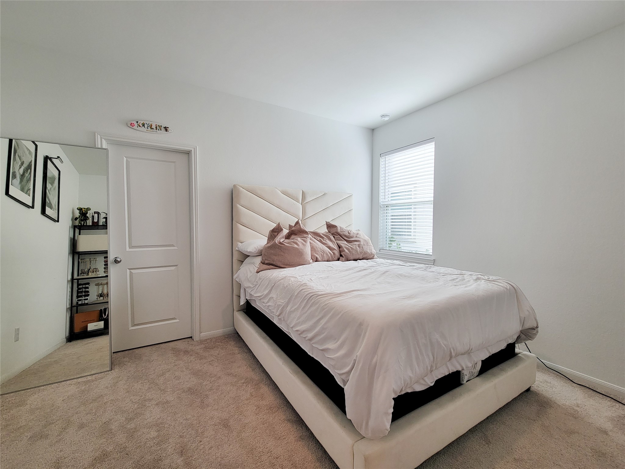 This bedroom features a cozy space with a plush carpet and a stylish, modern bed. It has a large window for natural light, a wall mirror, and a simple, clean design with white walls and minimalist decor.