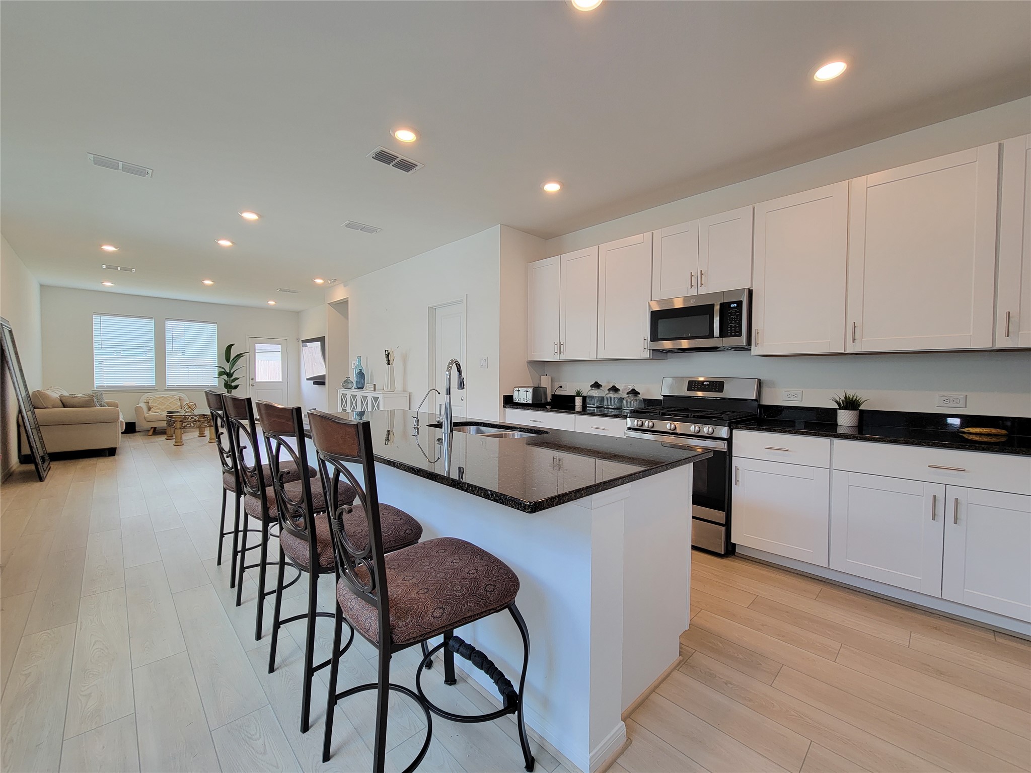 Modern, open-concept kitchen and living area. The kitchen features sleek white cabinetry, a large island with bar seating, and stainless steel appliances. The space is well-lit with recessed lighting, and the light wood flooring adds a warm touch. The living area in the background offers ample natural light from large windows.
