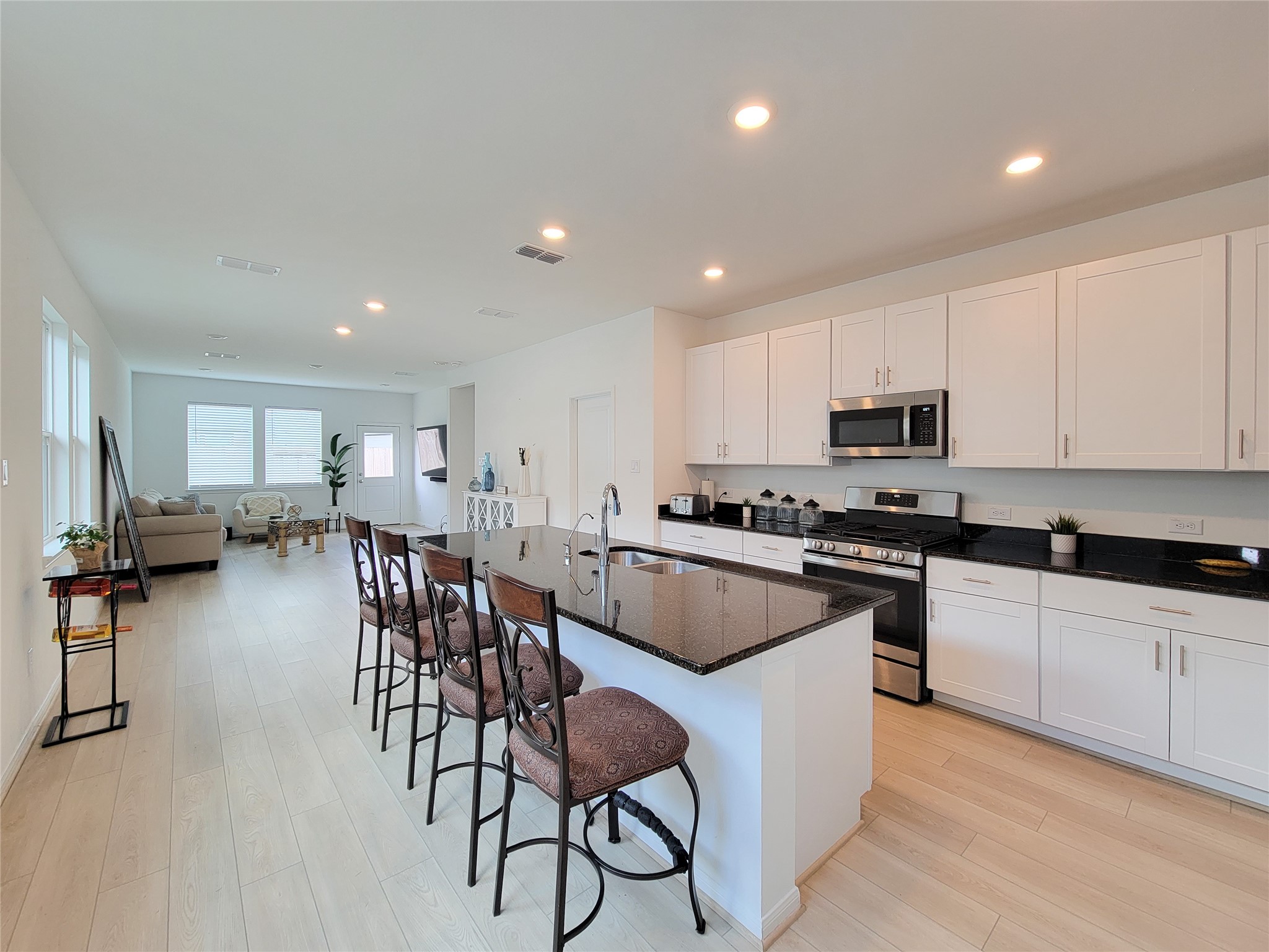 The kitchen features modern white cabinets, a sleek black granite countertop, and a central island with bar seating. The space is well-lit with recessed lighting, and the adjacent living area offers ample natural light, providing a warm and inviting atmosphere.