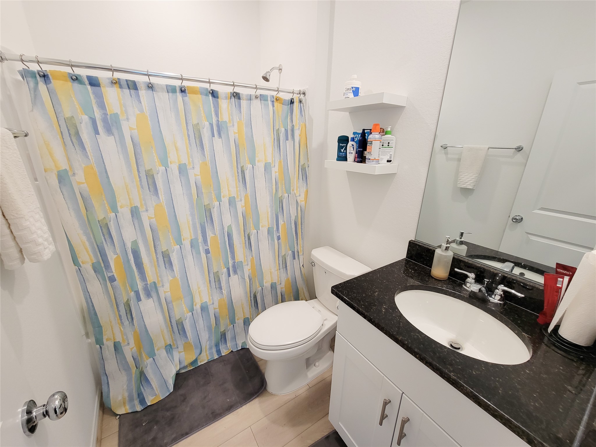 This bathroom features a modern design with a black granite countertop, white vanity, and a colorful shower curtain. It includes a standard toilet, shelves for storage, and a large mirror. The space is well-lit and has a clean, fresh appearance.