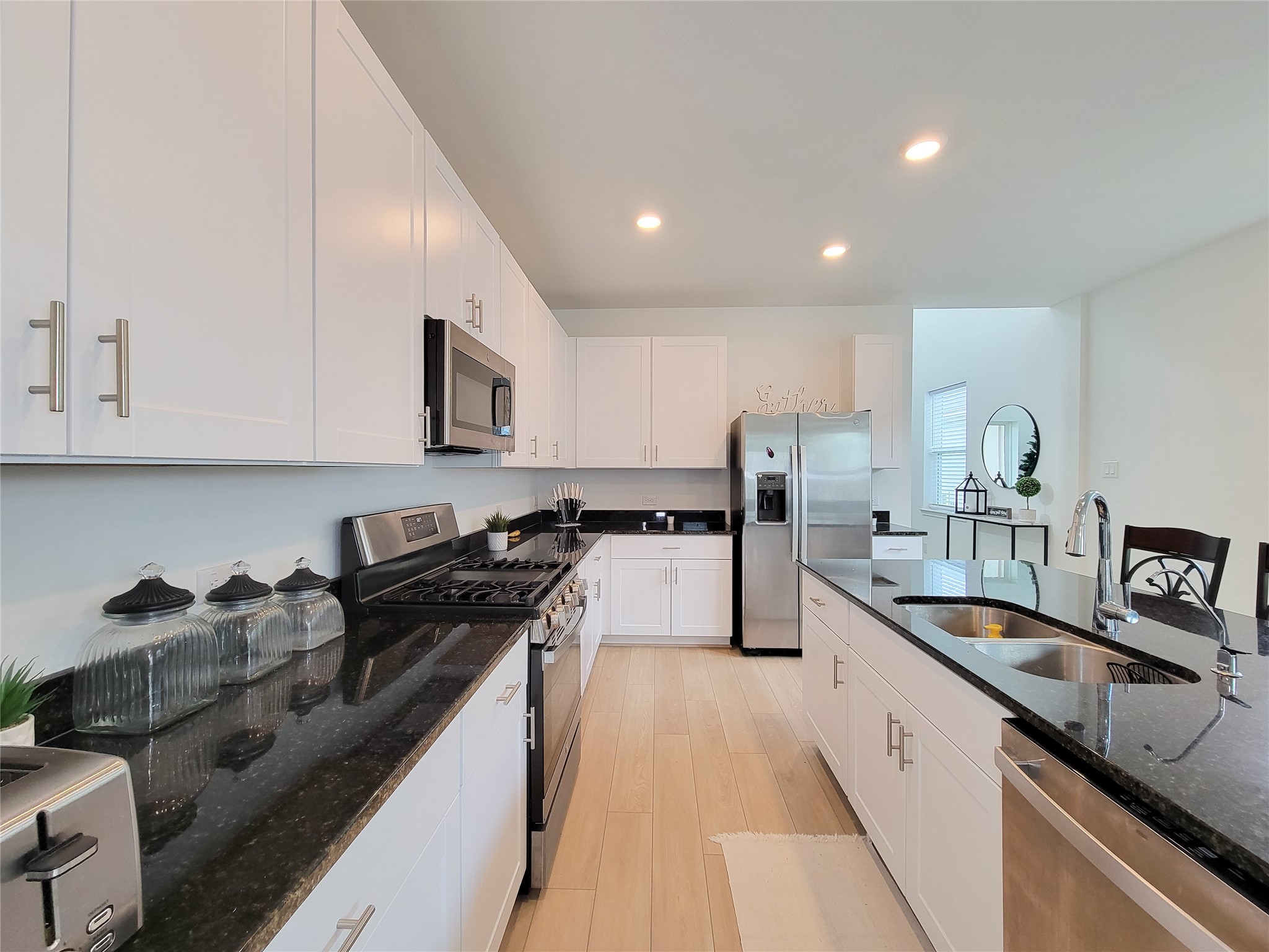 This modern kitchen features sleek white cabinetry, black granite countertops, and stainless steel appliances, including a double oven and fridge. The open layout and ample lighting create a bright, inviting space perfect for cooking and entertaining.