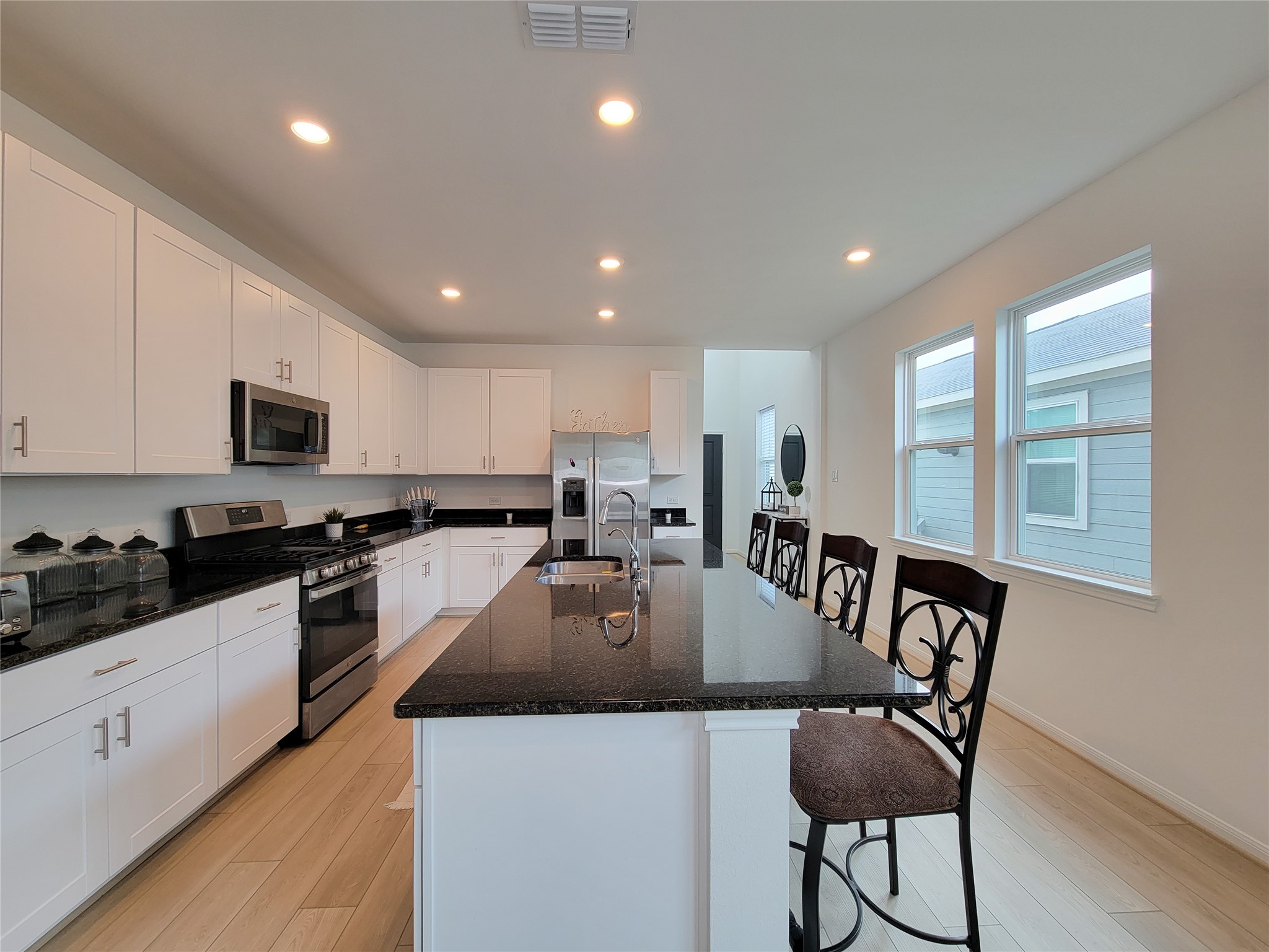 This kitchen features a modern design with white cabinetry, a sleek black granite countertop island, stainless steel appliances, and ample natural light from large windows. Perfect for entertaining and cooking.