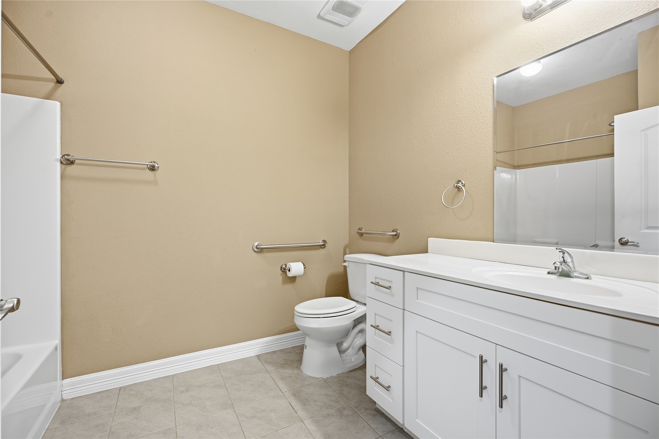 Located between the two additional bedrooms and easily accessed from the main living area, this bathroom features a white vanity with drawer and cabinet storage, a large mirror, and grab bars beside the toilet.