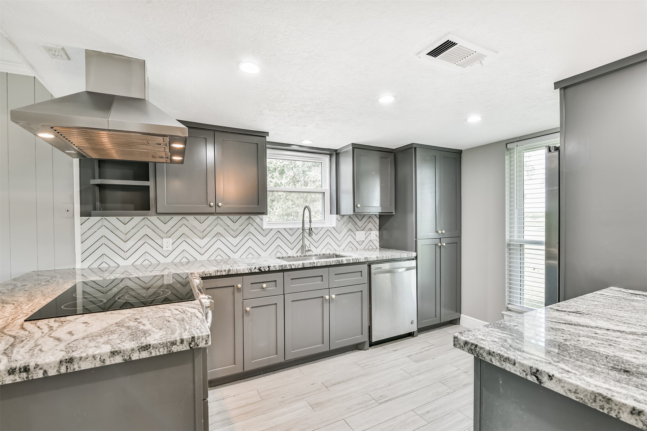 Modern kitchen featuring sleek gray cabinets, elegant herringbone backsplash, and stunning granite countertops. Abundant natural light and top-of-the-line appliances create a perfect space for culinary creativity.