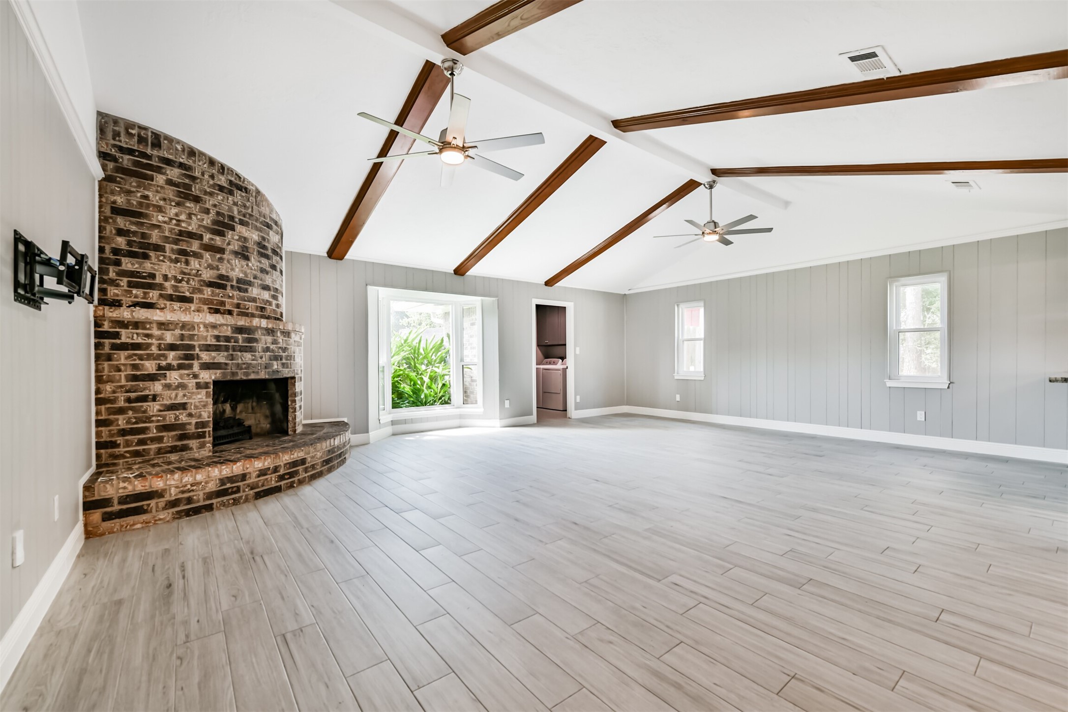 Spacious living area featuring vaulted ceilings with elegant wood beams, a stunning brick fireplace, and abundant natural light from large windows. Perfect for relaxing or entertaining.
