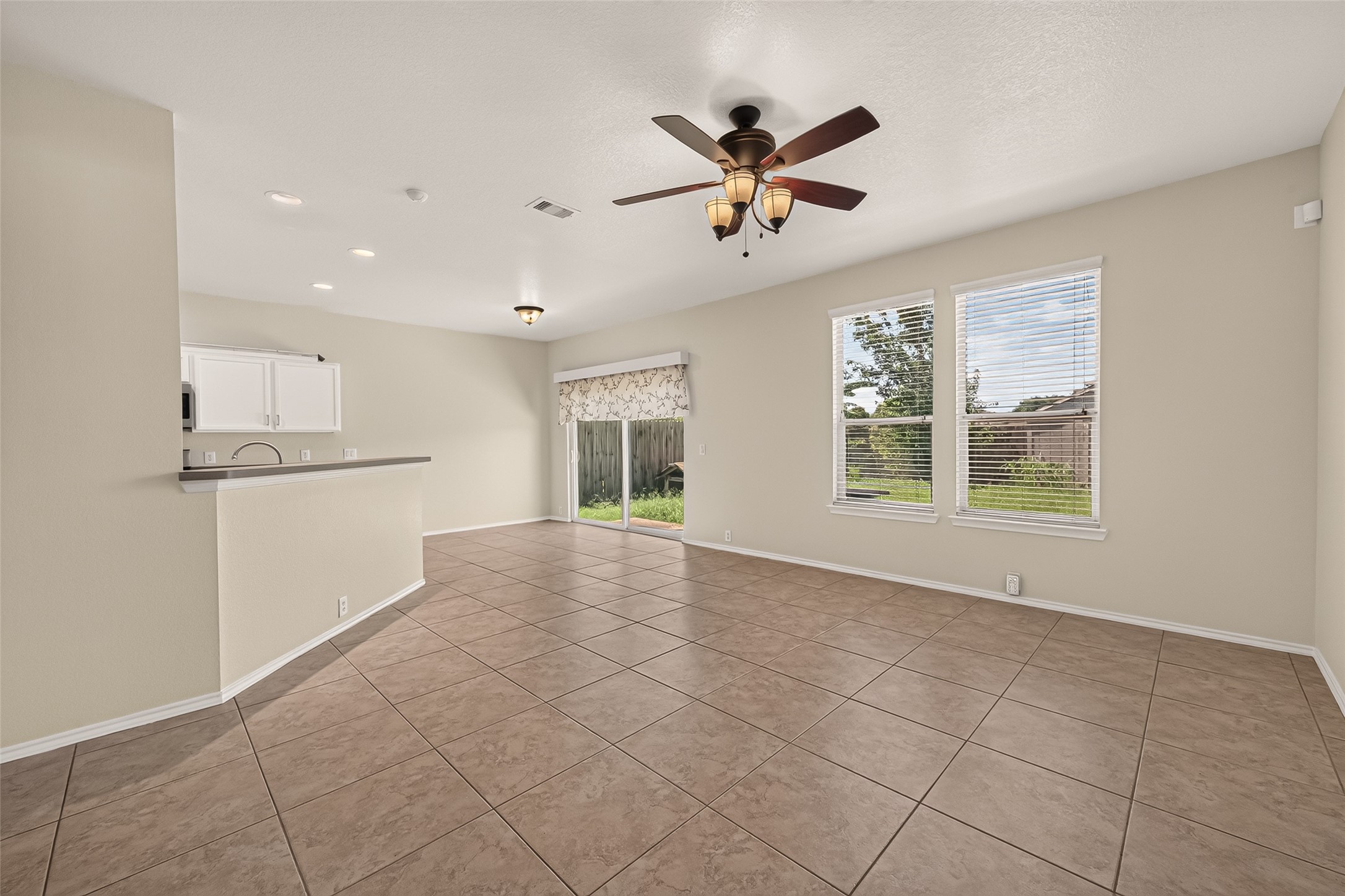 Bright open living space with large windows, tiled flooring, and a ceiling fan. The room connects to a kitchen with a breakfast bar, and a sliding glass door leads to the backyard.