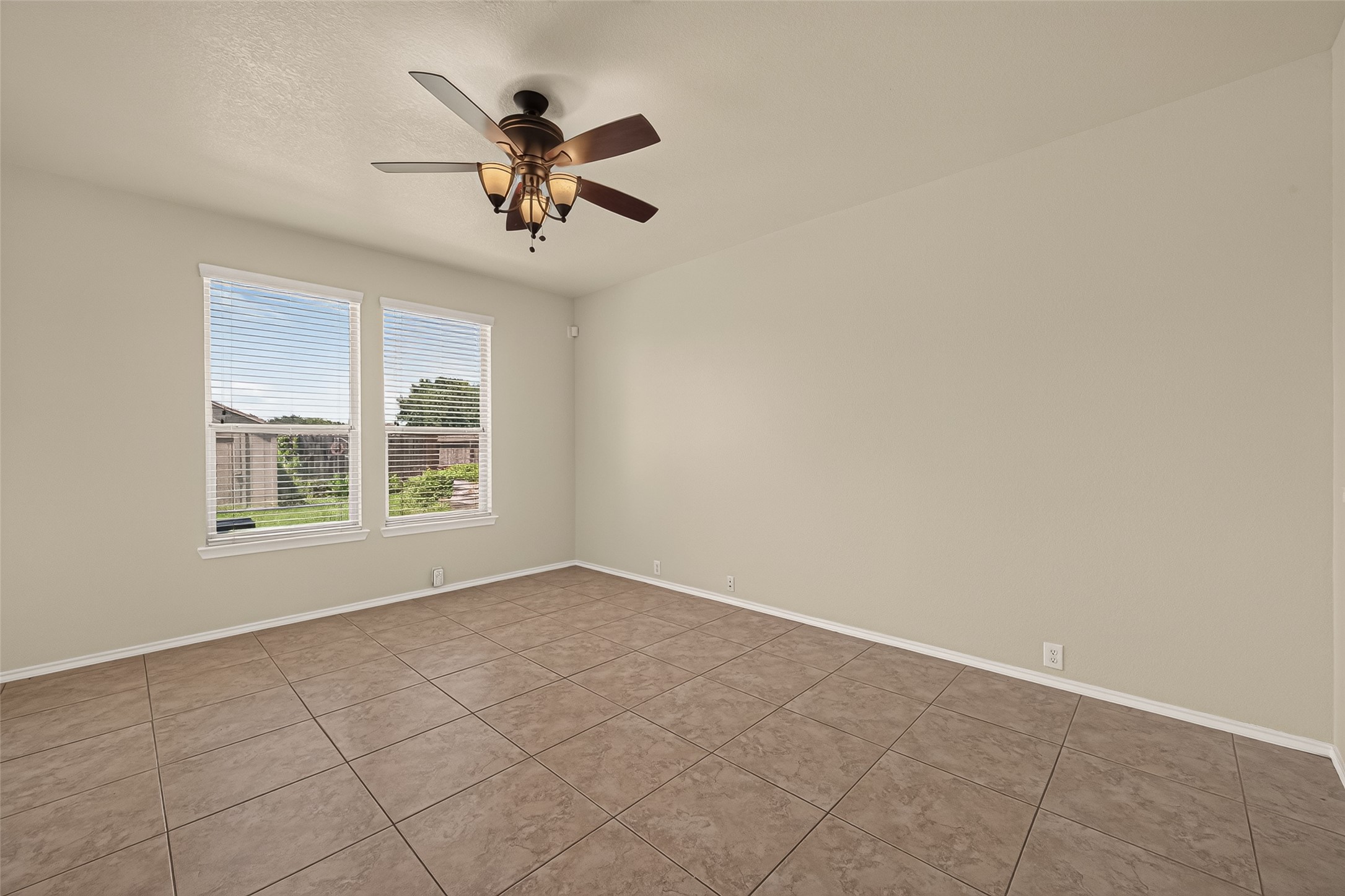 This room features neutral walls, tile flooring, and a ceiling fan with lights. Two large windows provide natural light and a view of the outdoors, creating a bright and airy space.