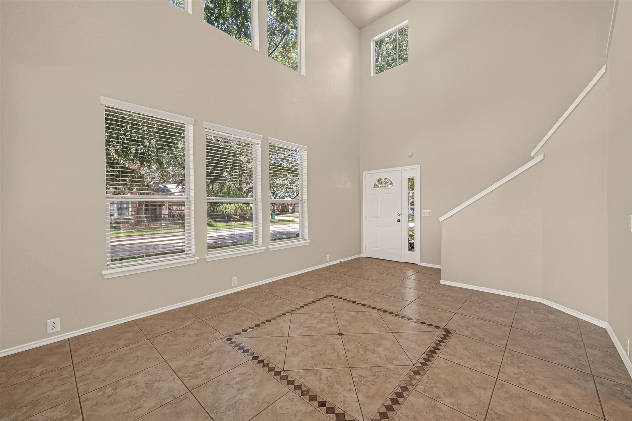 This photo showcases a bright, spacious room with high ceilings and large windows that let in plenty of natural light. The room features neutral-colored walls and tiled flooring with a decorative inlay. A white front door and a staircase leading upwards add to the inviting atmosphere.