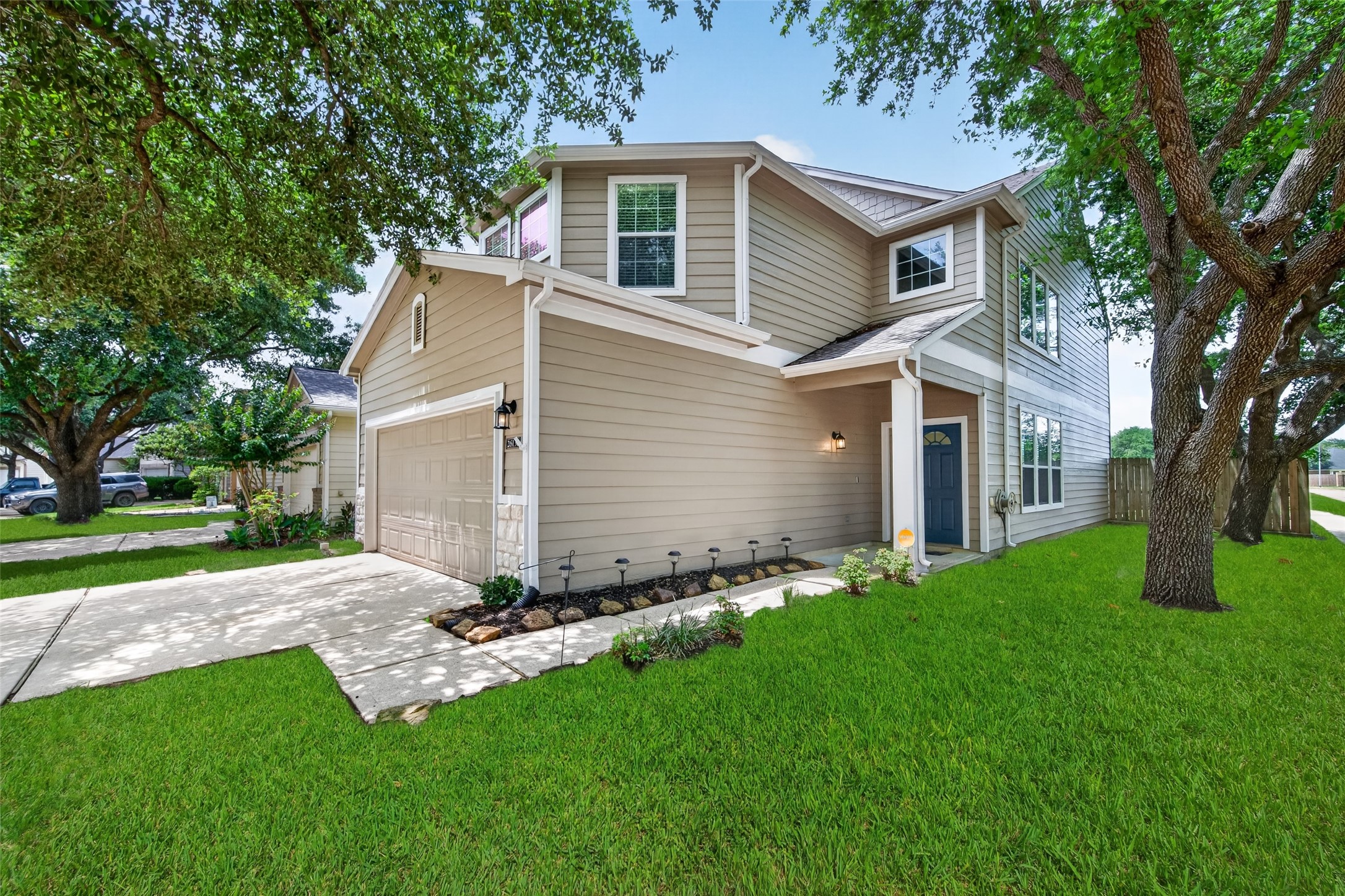 This photo shows a charming two-story home with beige siding and white trim. It features a double garage, a well-maintained front lawn, and mature trees providing shade. The entrance has a welcoming porch with a blue door, giving the home a cozy and inviting feel.
