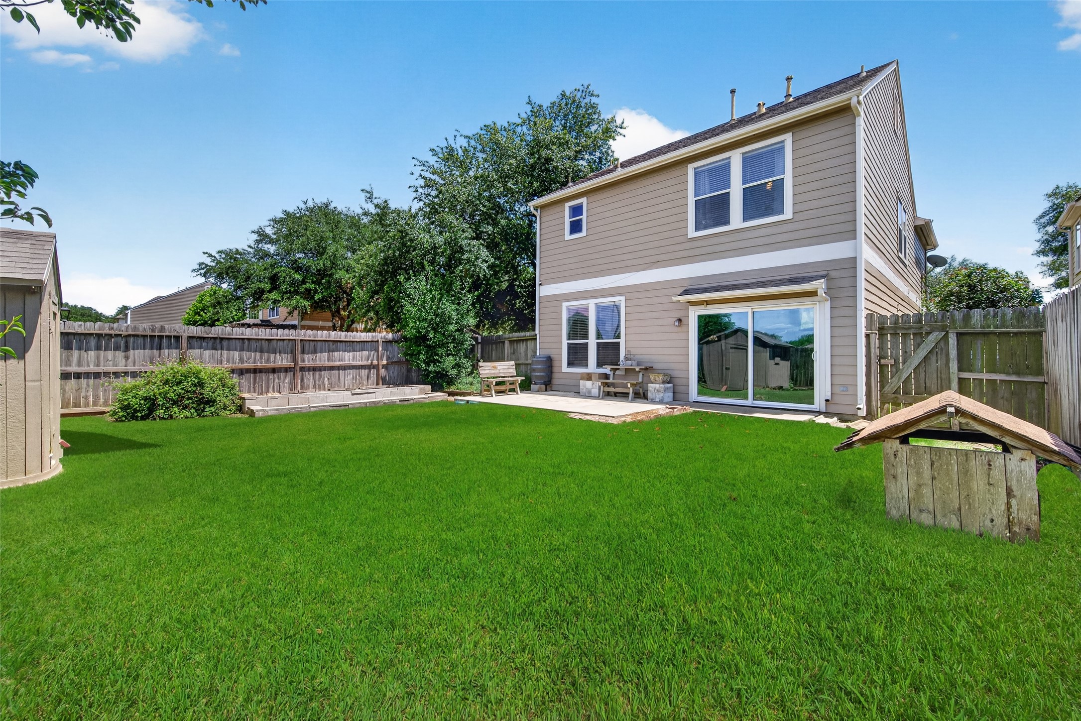 This backyard features a spacious, grassy area with a wooden fence for privacy. There's a concrete patio with seating, perfect for outdoor relaxation or entertaining. The exterior of the house is a modern two-story design with large windows that offer plenty of natural light.