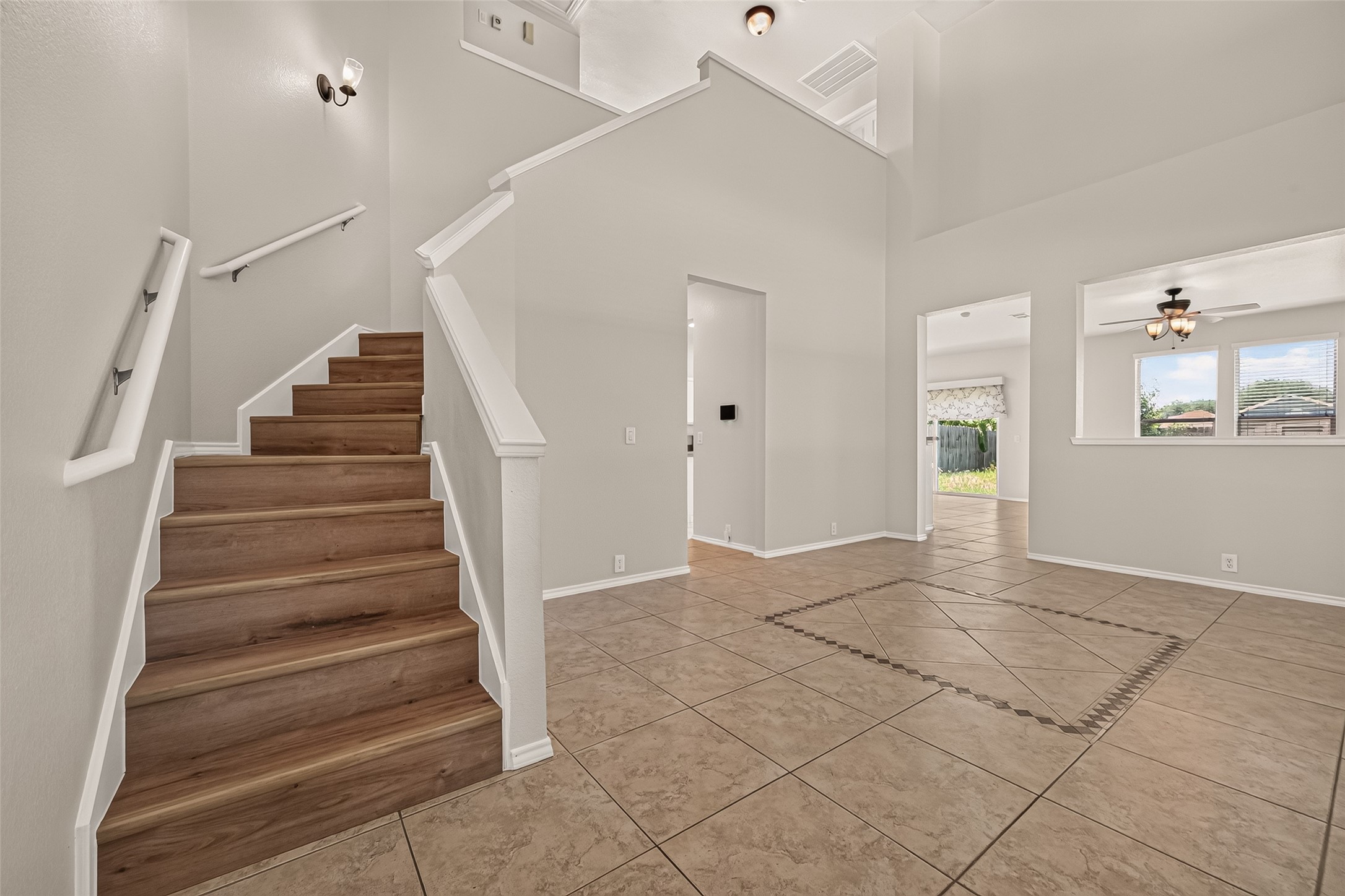 This photo showcases a bright, open entryway with neutral walls and tiled flooring. A wooden staircase leads upstairs, and there are open doorways providing a view to additional rooms, including one with large windows and a ceiling fan. The space feels modern and welcoming.