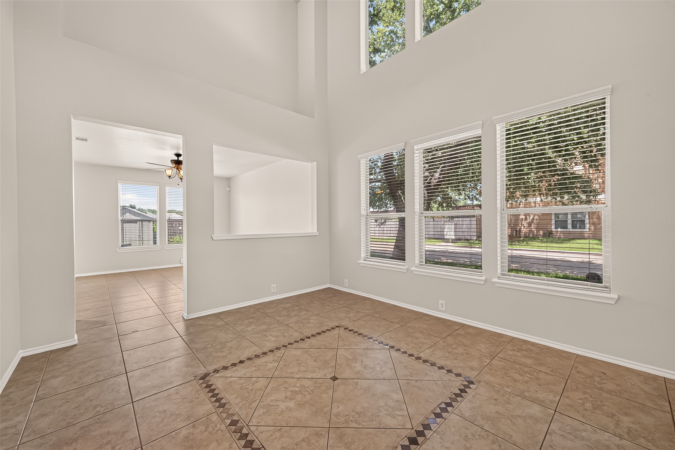 This spacious room features high ceilings and large windows that let in plenty of natural light. The tile flooring includes a decorative inlay, and there's an open view into an adjacent room with a ceiling fan.