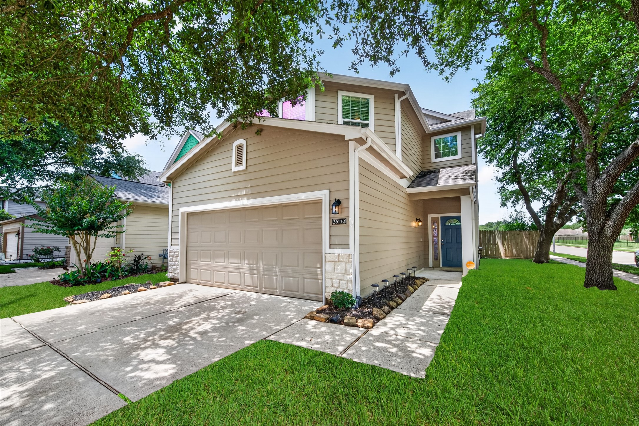 This charming two-story home features a beige exterior with stone accents, a two-car garage, and a welcoming front entrance. It's surrounded by lush green lawns and mature trees, providing a serene and inviting atmosphere.