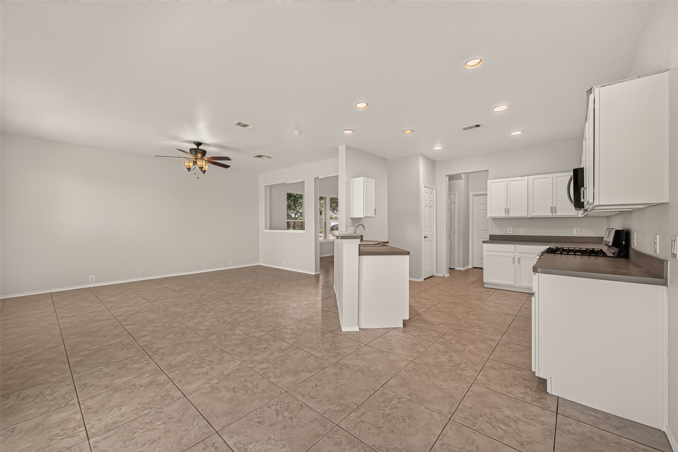 This photo showcases a spacious open-concept living area with tiled floors, a modern kitchen featuring white cabinetry, and a ceiling fan. The area is well-lit with recessed lighting and offers ample space for versatile furniture arrangements.