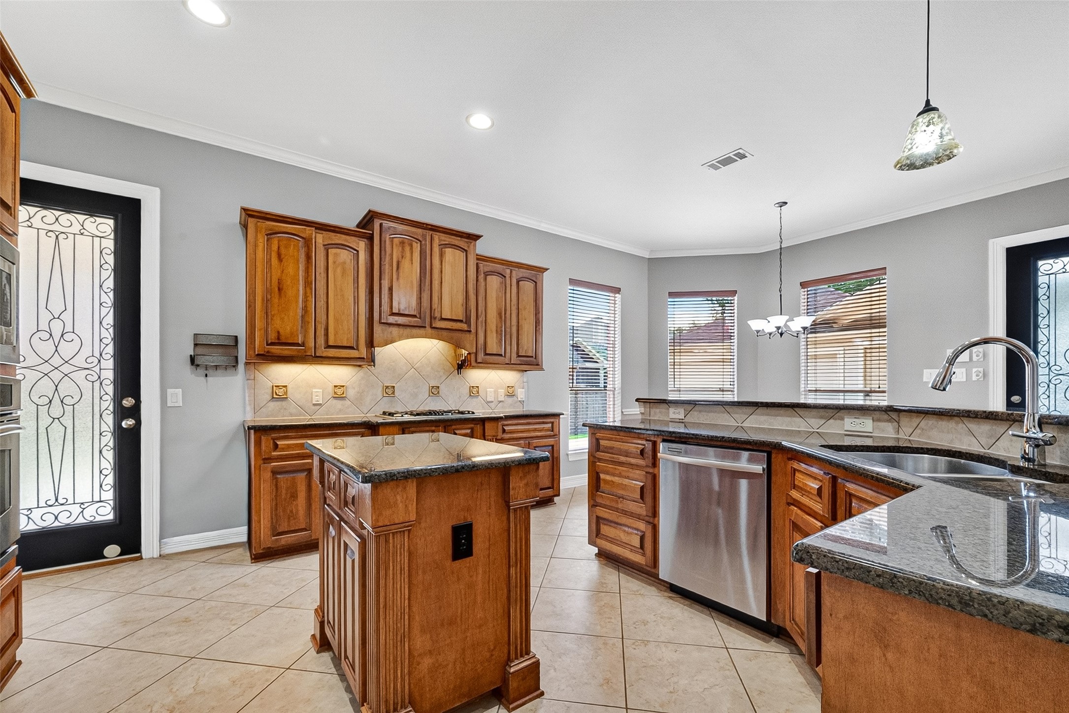 This inviting kitchen features large windows for natural light, a stylish chandelier, and a decorative glass door leading outside. The space is enhanced by neutral tile flooring and a wooden dining set, creating a warm, functional atmosphere.