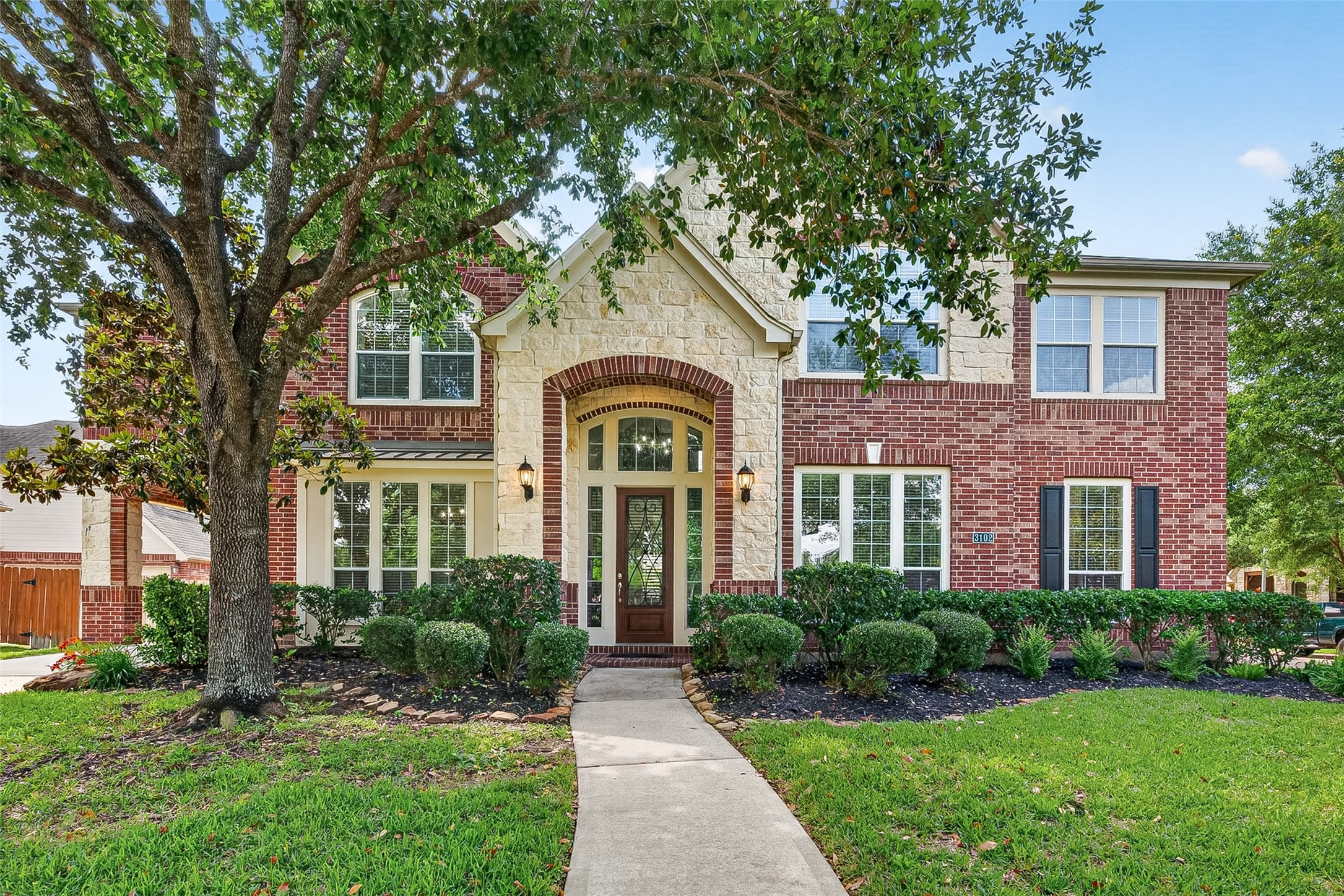This 6-bed home is a two-story adorned with brick and stone with a welcoming front facade and lush landscaping. It features a spacious covered driveway leading to a garage, complemented by mature trees and a well-maintained lawn.