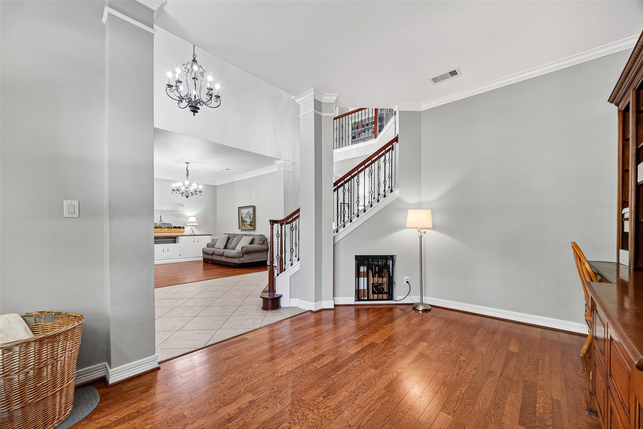 Bright, elegant entryway with hardwood floors and a stylish staircase. The space opens into a living area with tiled flooring and decorative chandeliers.