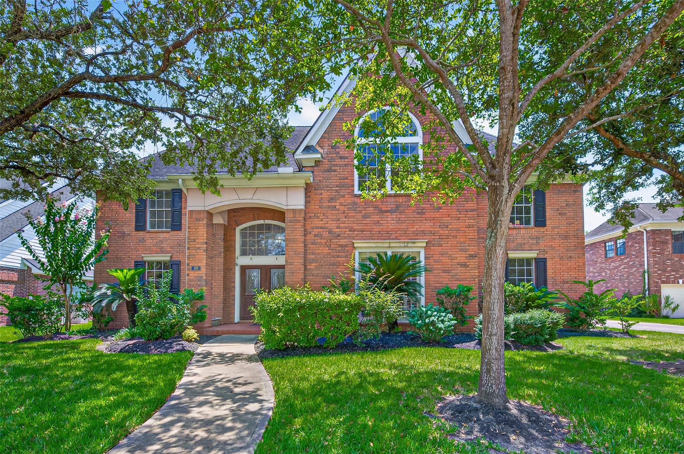 Elegant two-story brick home with charming arched entryway, surrounded by lush landscaping and shaded by mature trees.