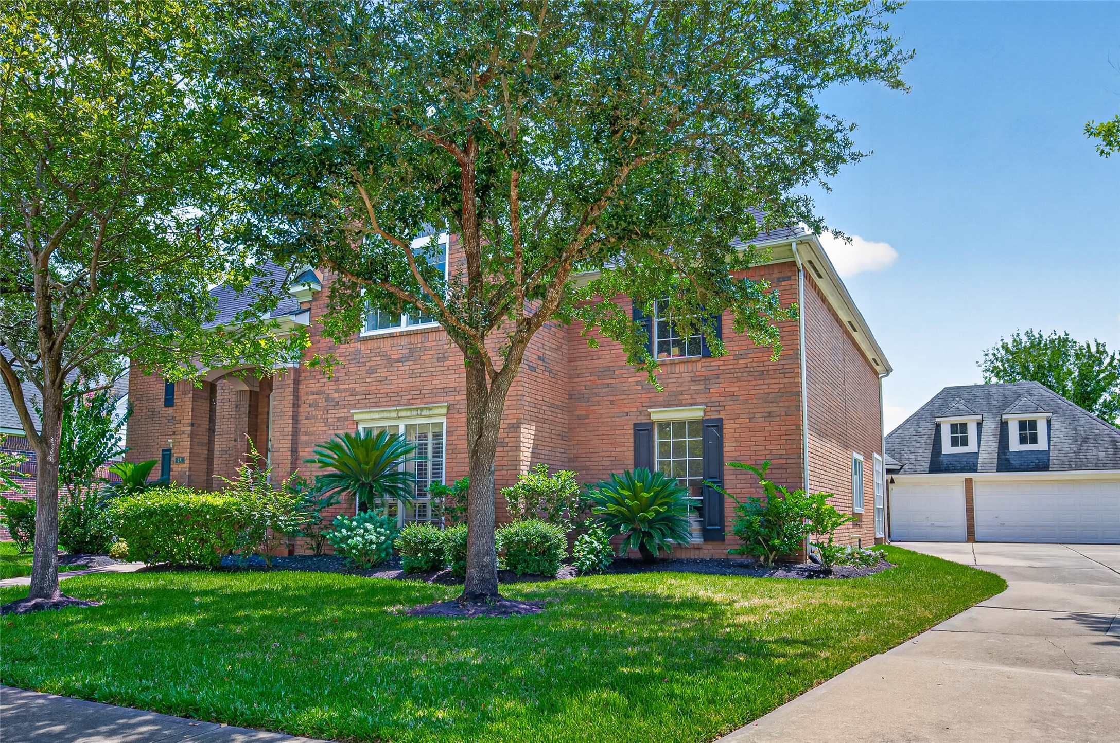 Charming brick home with lush landscaping and mature trees, offering great curb appeal and a spacious driveway leading to a detached garage.
