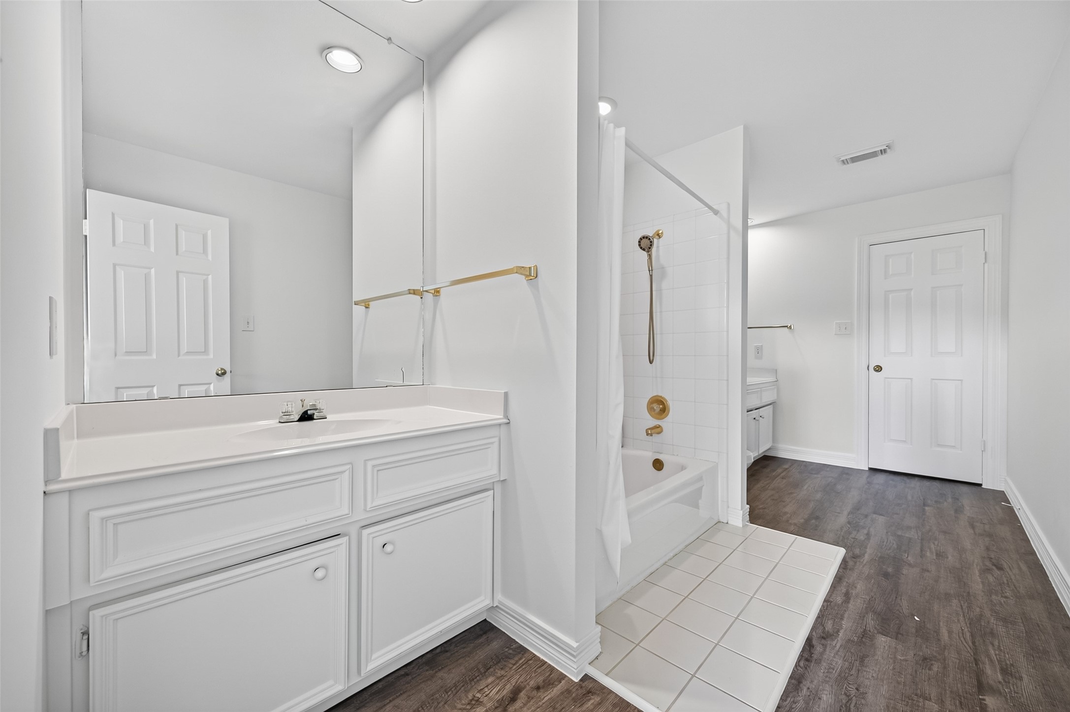 Bright and spacious bathroom featuring a large vanity with ample cabinets, a classic white tub with gold fixtures, and elegant dark flooring.