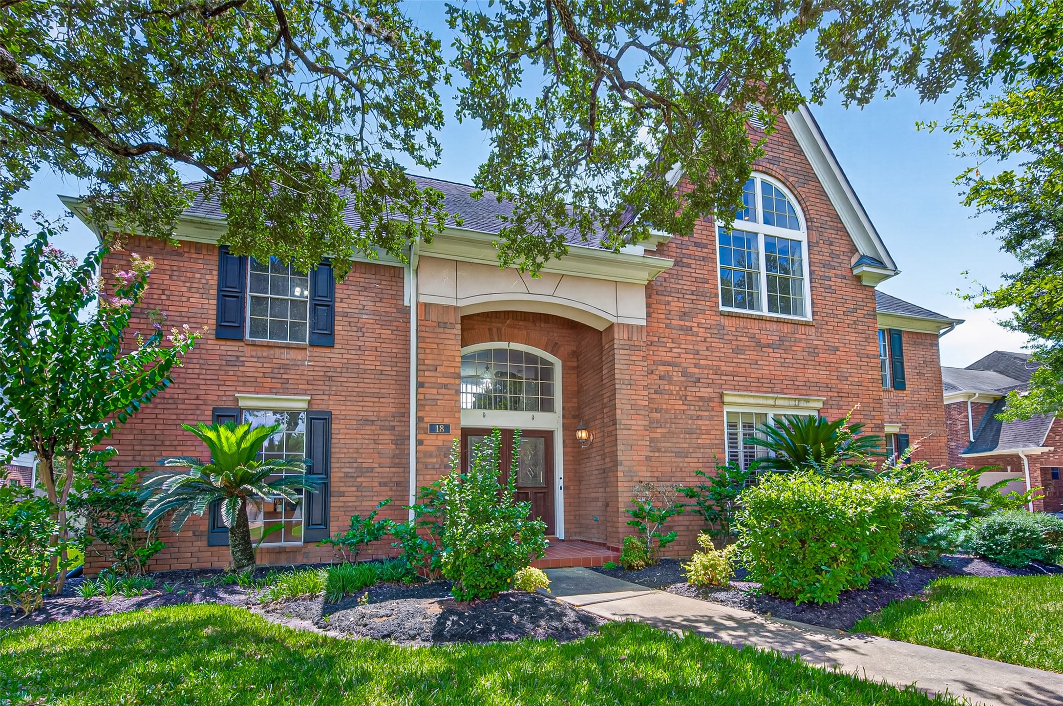 Stately brick home with elegant arched windows, lush landscaping, and a welcoming entrance framed by mature trees.