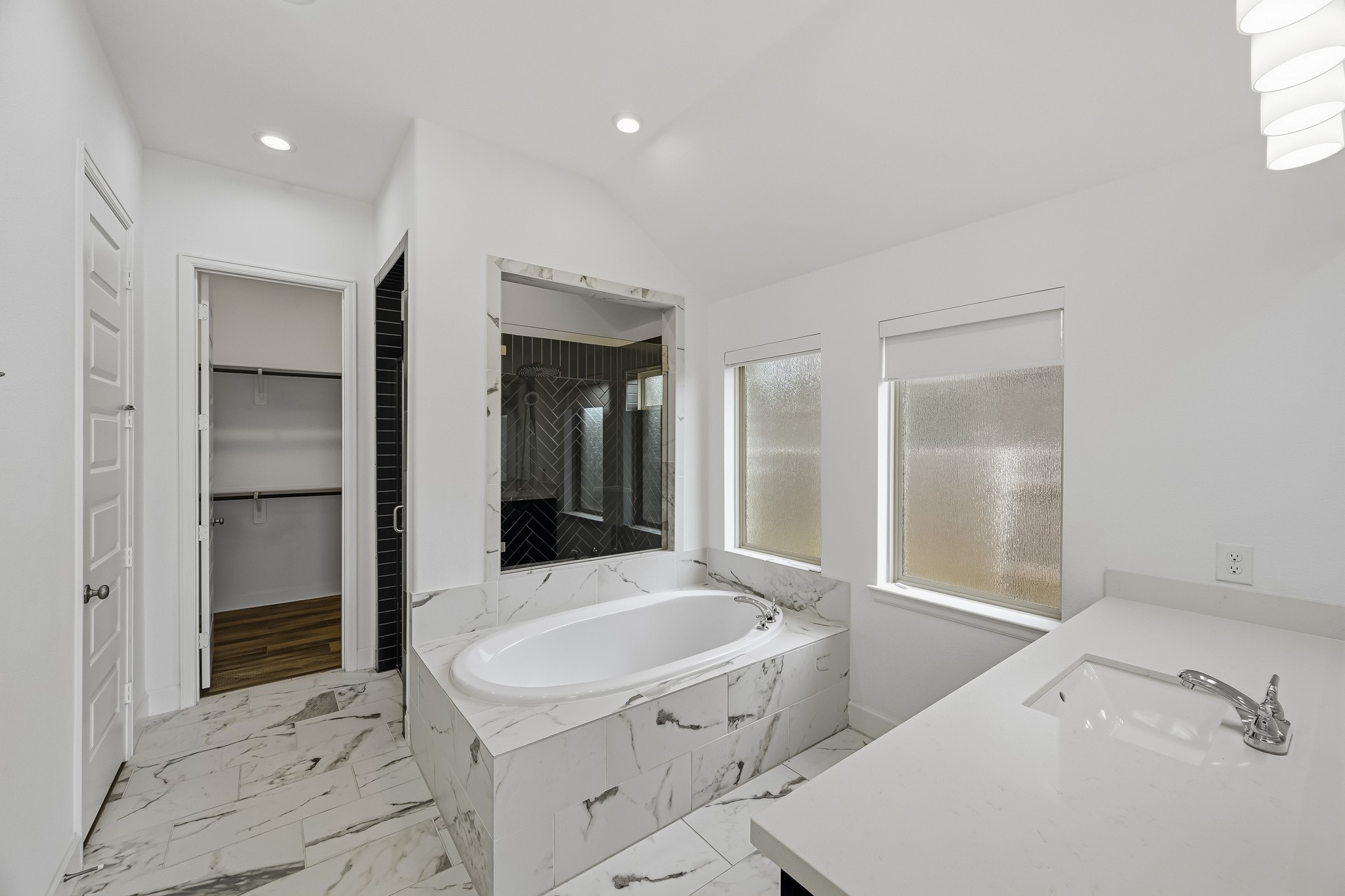 Welcome to an expansive master bath where vaulted ceilings and recessed lighting create an open, luminous sanctuary. Polished marble-style tile extends beneath a built-in soaking tub, filled with soft natural light from large privacy windows.