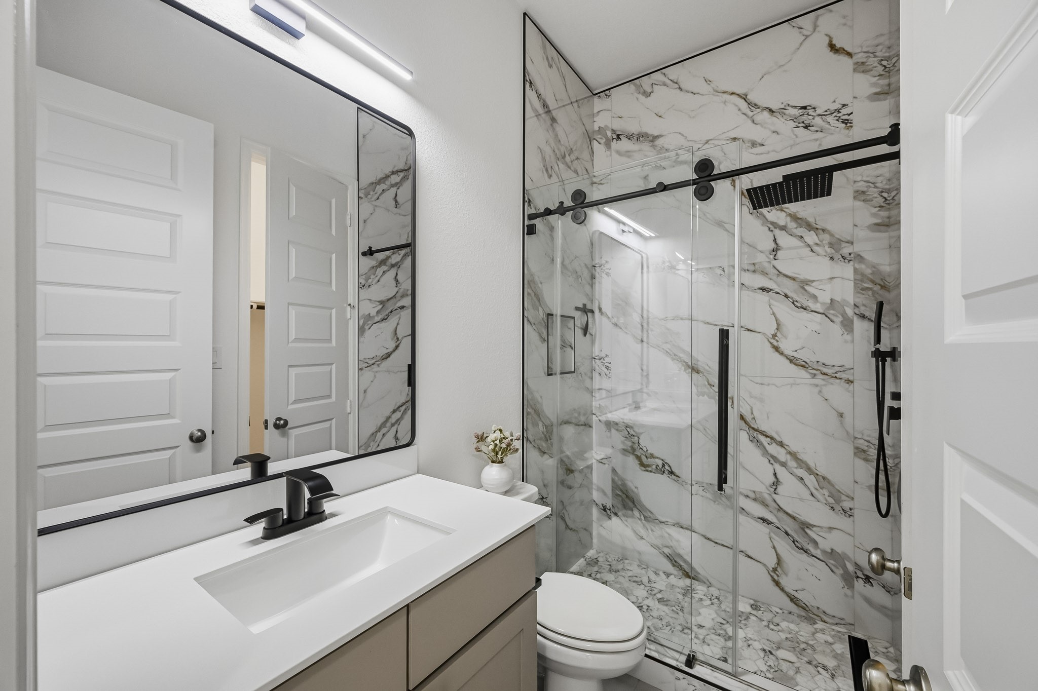 Sophisticated finishes define this well-proportioned secondary bathroom retreat. Highlights include premium marble-pattern wall surfaces extending through the glass-enclosed shower, streamlined hardware, and an expansive wall mirror.