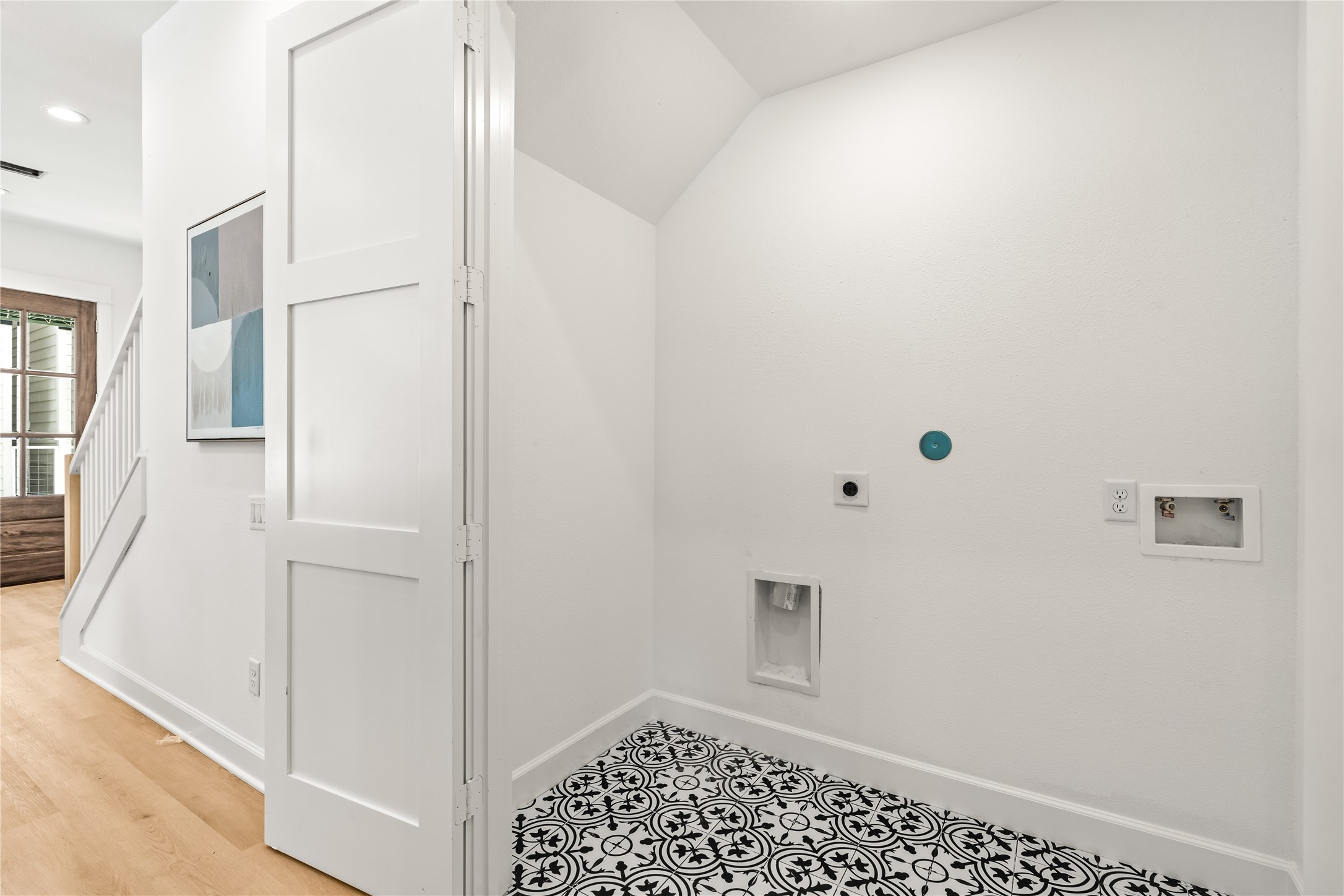 A dedicated laundry closet nestled under the stairs, showcasing graphic black-and-white patterned floor tile, standard utility hookups, and a white bifold privacy door.
