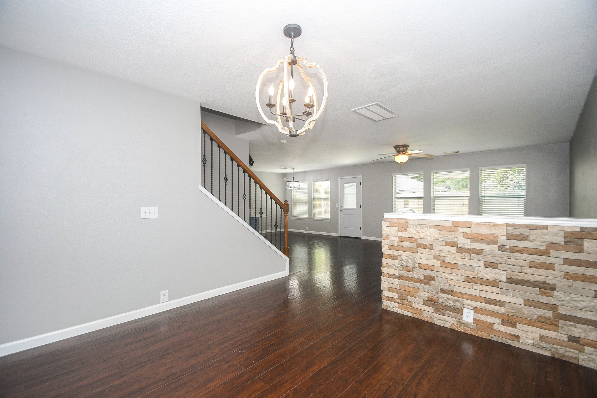 Spacious open-concept living area features elegant laminate wood floors, a modern light fixture, and a stylish stone accent wall, leading to a staircase with wrought iron railings.