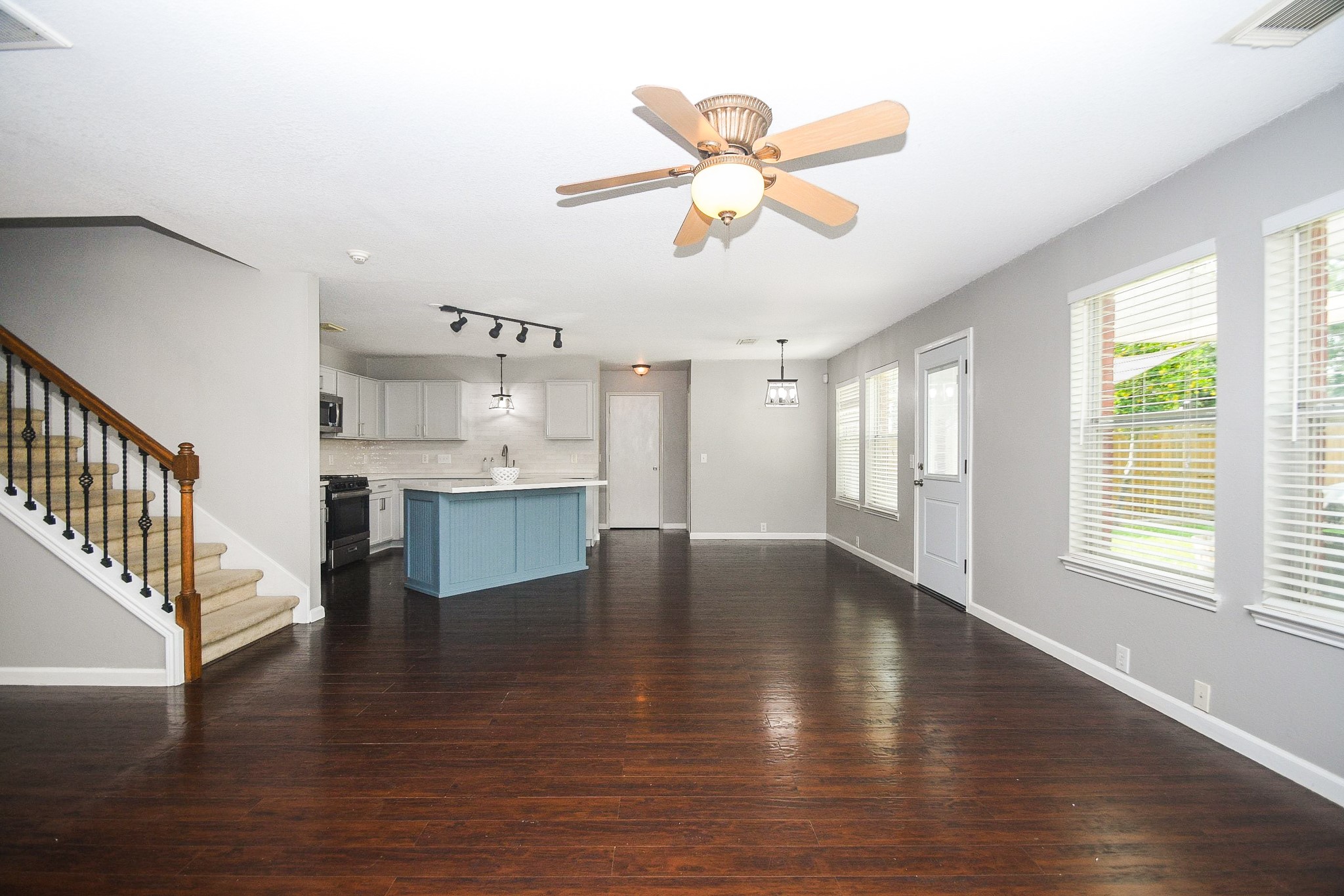Spacious open-concept living area with rich hardwood floors, modern ceiling fan, and abundant natural light from large windows. The kitchen features a stylish blue island, sleek white cabinetry, and stainless steel appliances. Adjacent is a cozy dining area, while a carpeted staircase leads to the upper level.