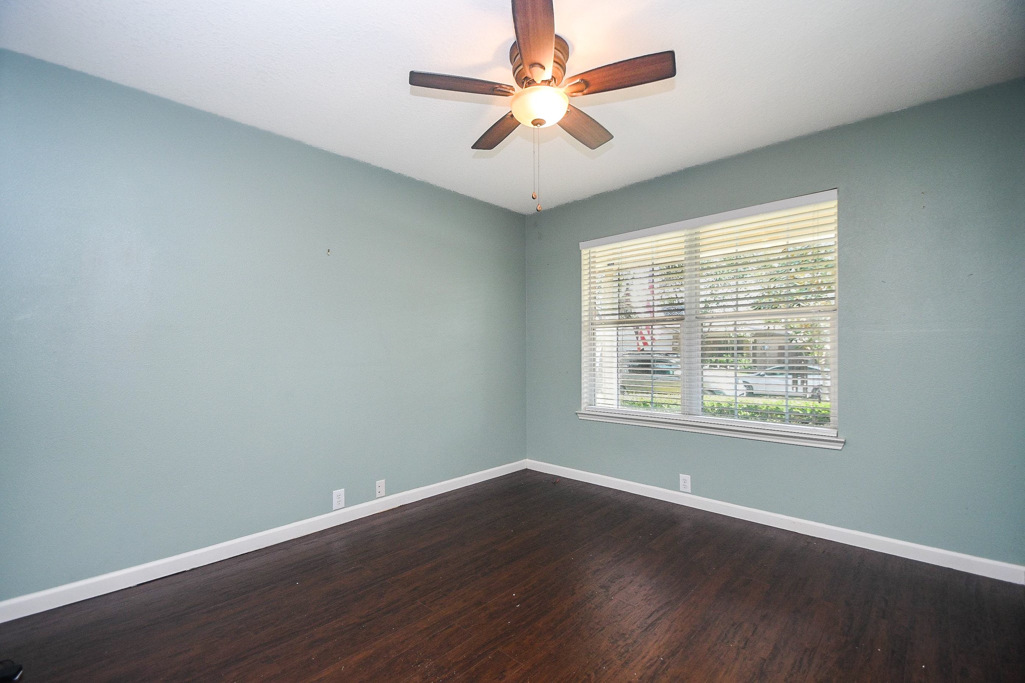 Spacious home office/study with rich, laminate wood flooring, a soothing blue accent wall, and ample natural light from a large window, complemented by a modern ceiling fan. Spacious home office/study with rich, laminate wood flooring, a soothing blue accent wall, and ample natural light from a large window, complemented by a modern ceiling fan.