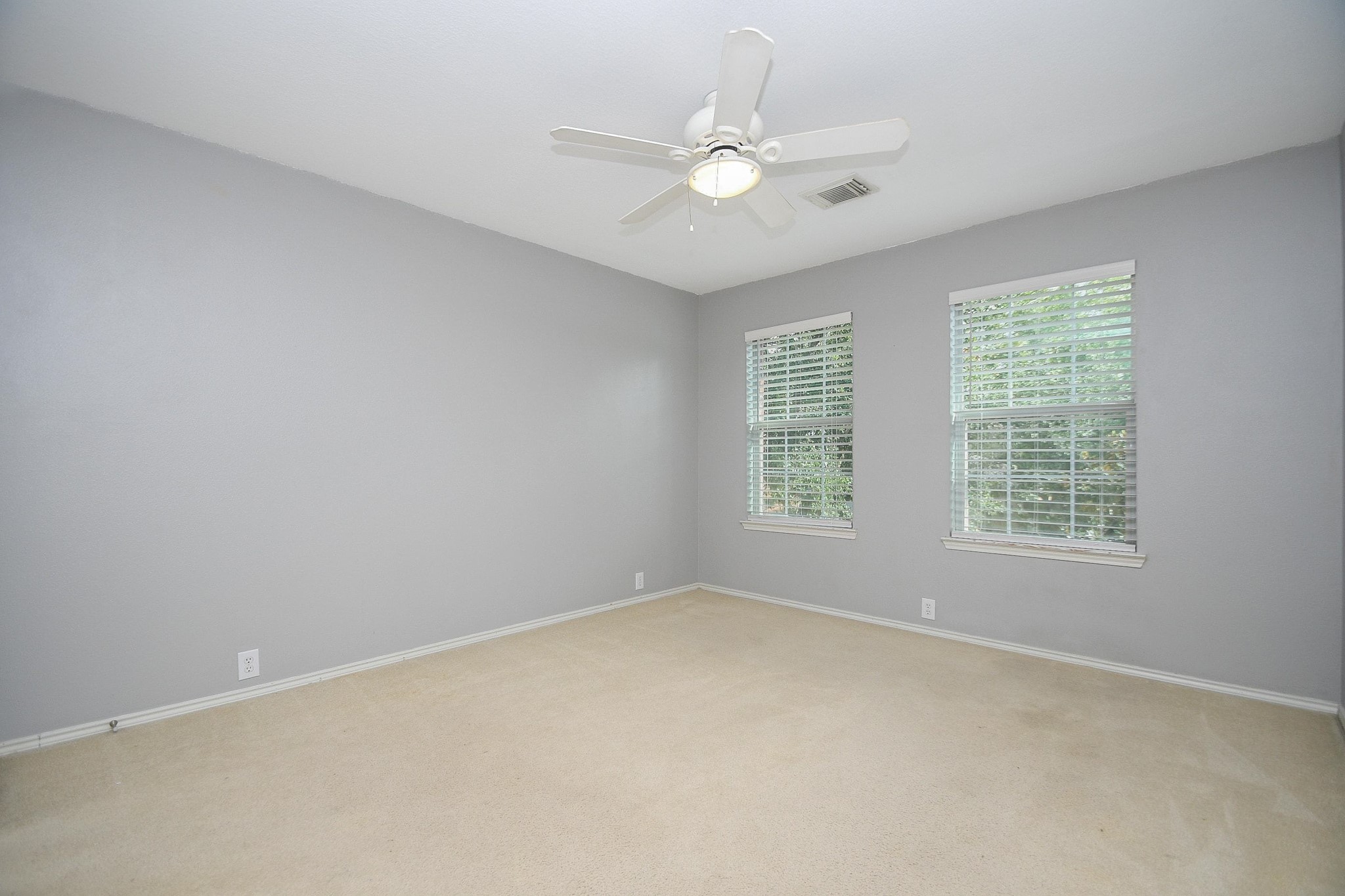 Spacious bedroom with soft gray walls, plush carpeting, and ample natural light from two large windows, complete with a ceiling fan for comfort.