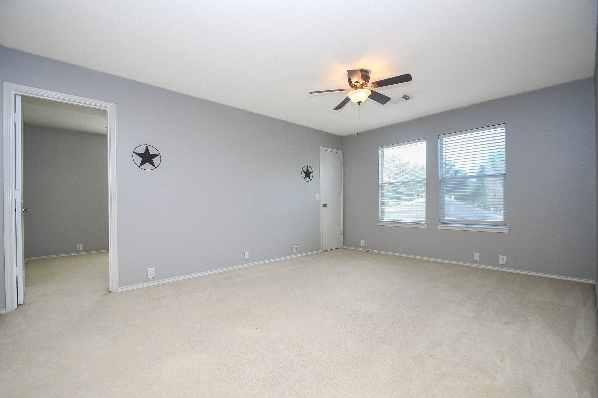 Spacious bedroom with soft carpet, neutral gray walls, and ample natural light from two large windows, complemented by a ceiling fan for comfort.