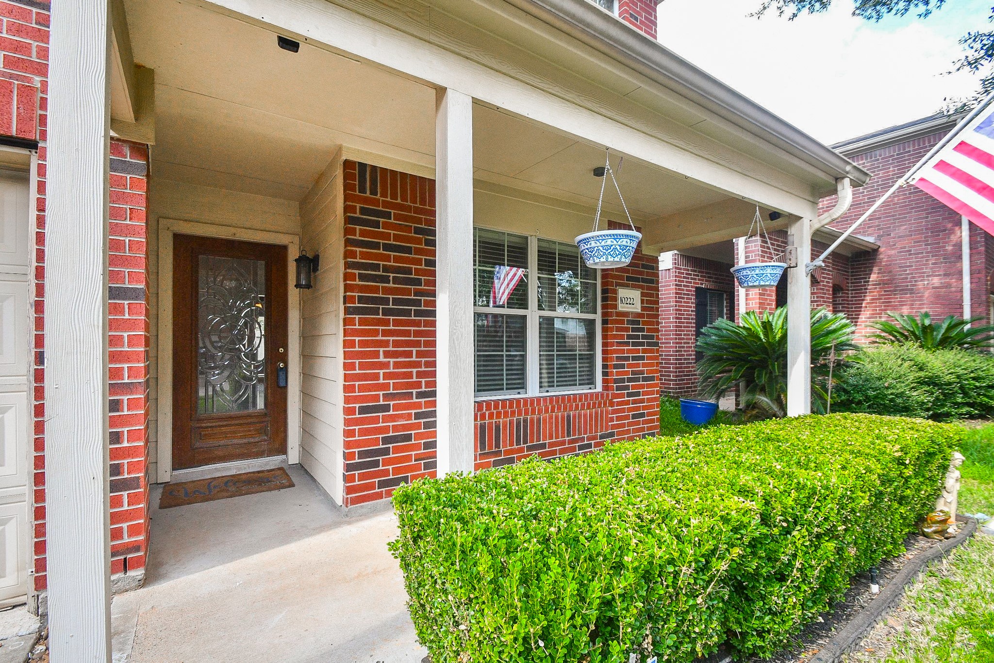 Charming brick home with a welcoming front porch, decorative glass door, and lush greenery, perfect for relaxing outdoors.