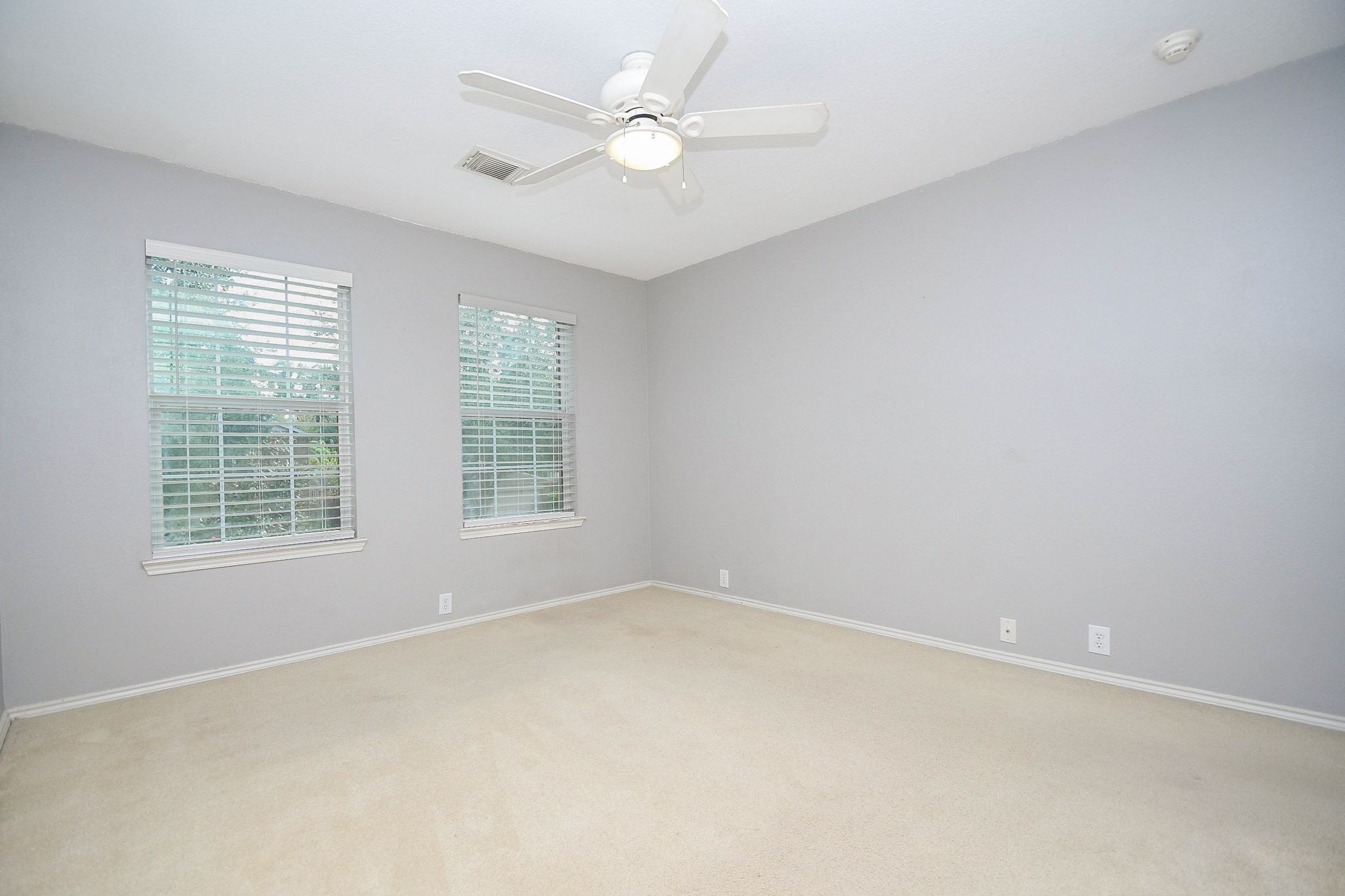 Spacious room with soft gray walls, plush carpeting, and ample natural light from two large windows, complete with a ceiling fan for comfort.